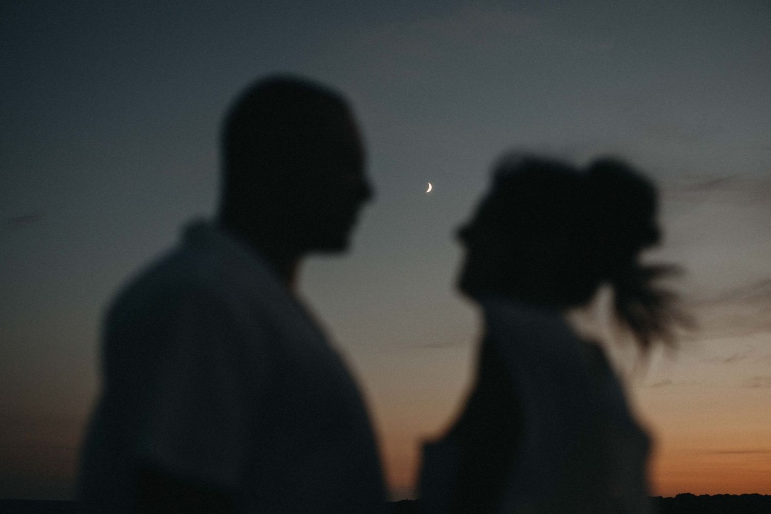 Romantic silhouette of a couple facing each other under a crescent moon at dusk.