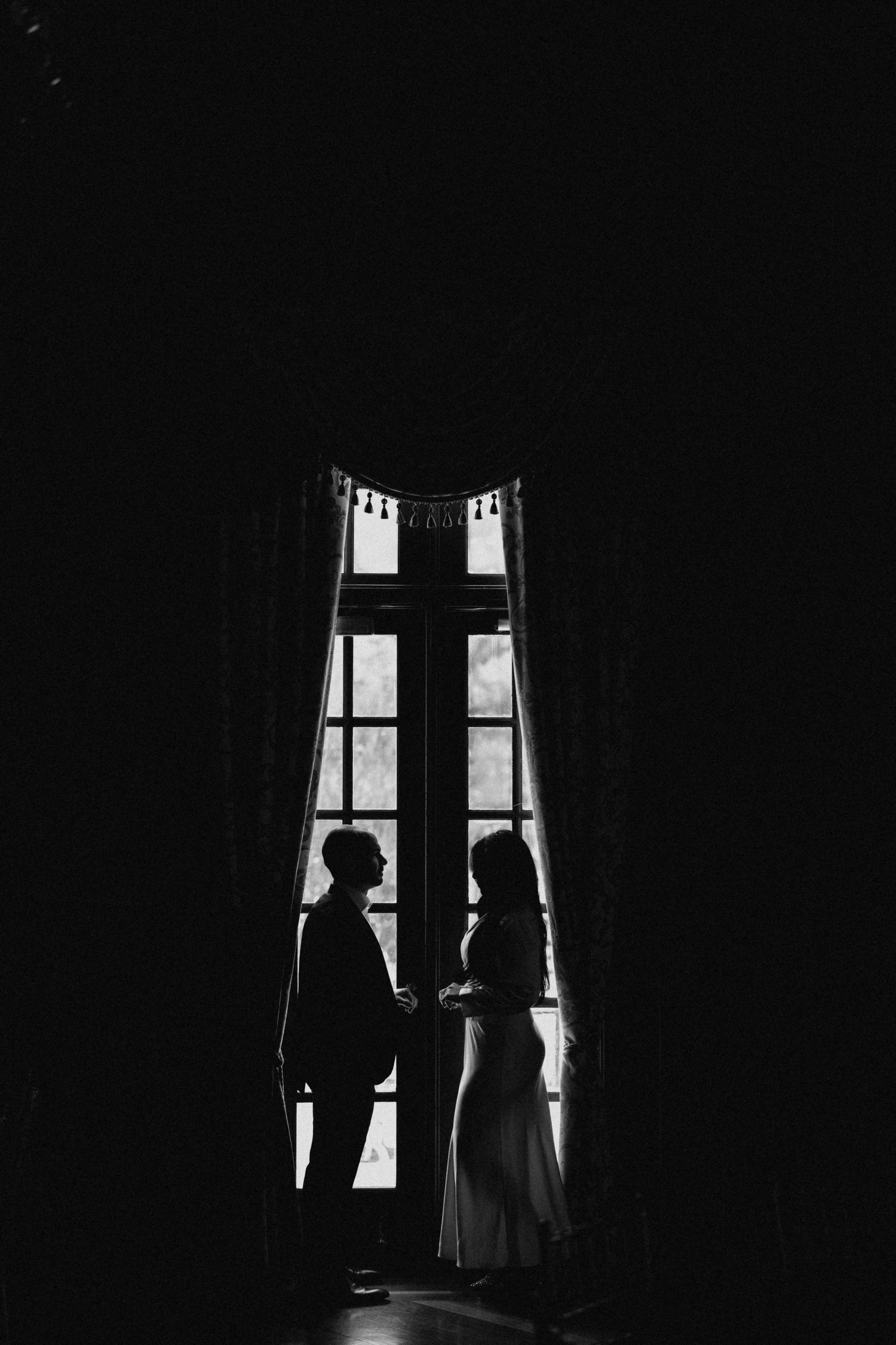 Black and white silhouette of a couple holding hands before a large ornate window indoors.