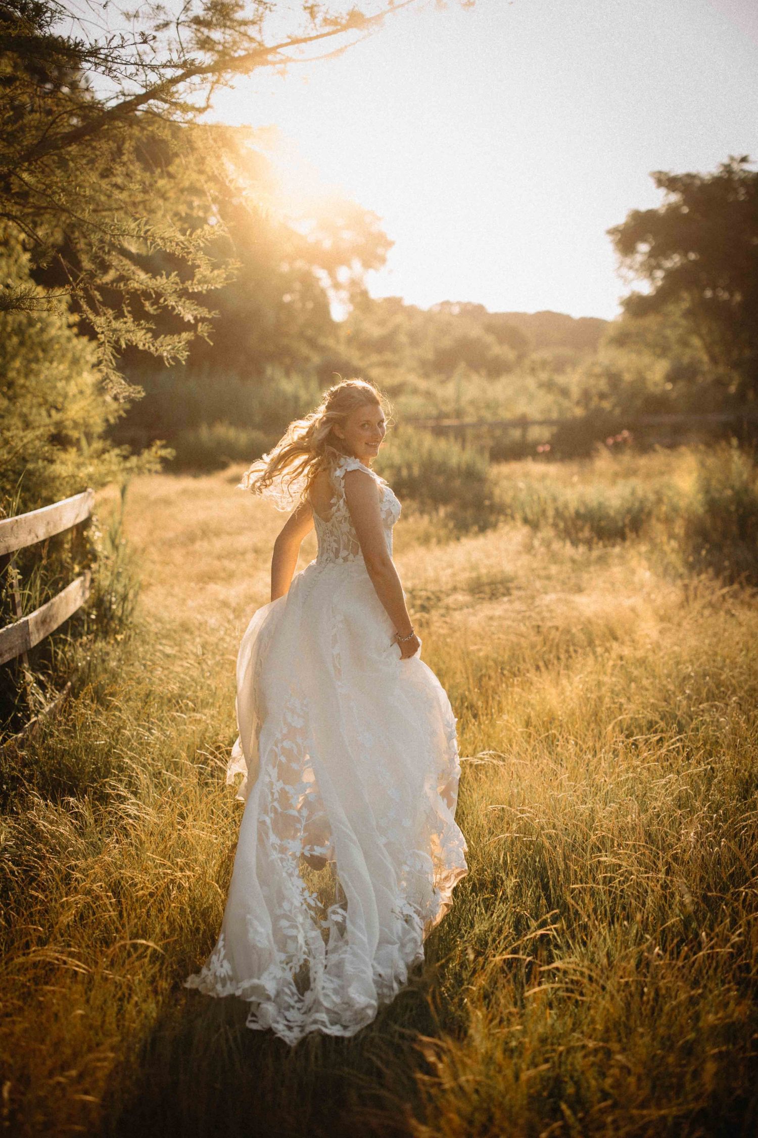 Bride in lace A-line wedding gown walks through golden sunlit meadow at sunset near a wooden fence.