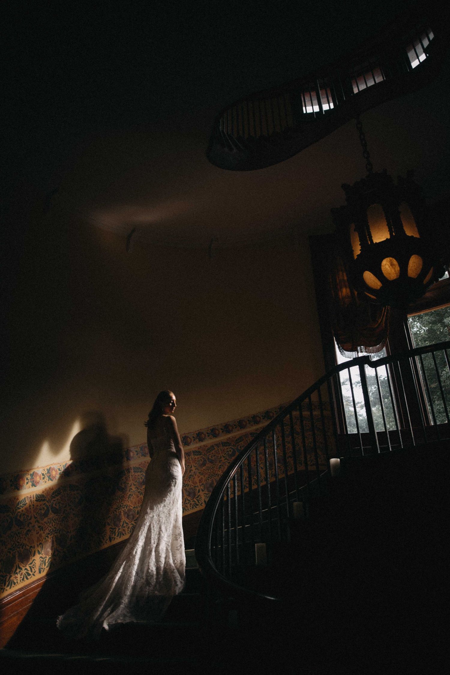 Bride in lace wedding gown standing on a grand staircase illuminated by a vintage lantern in a dark venue.