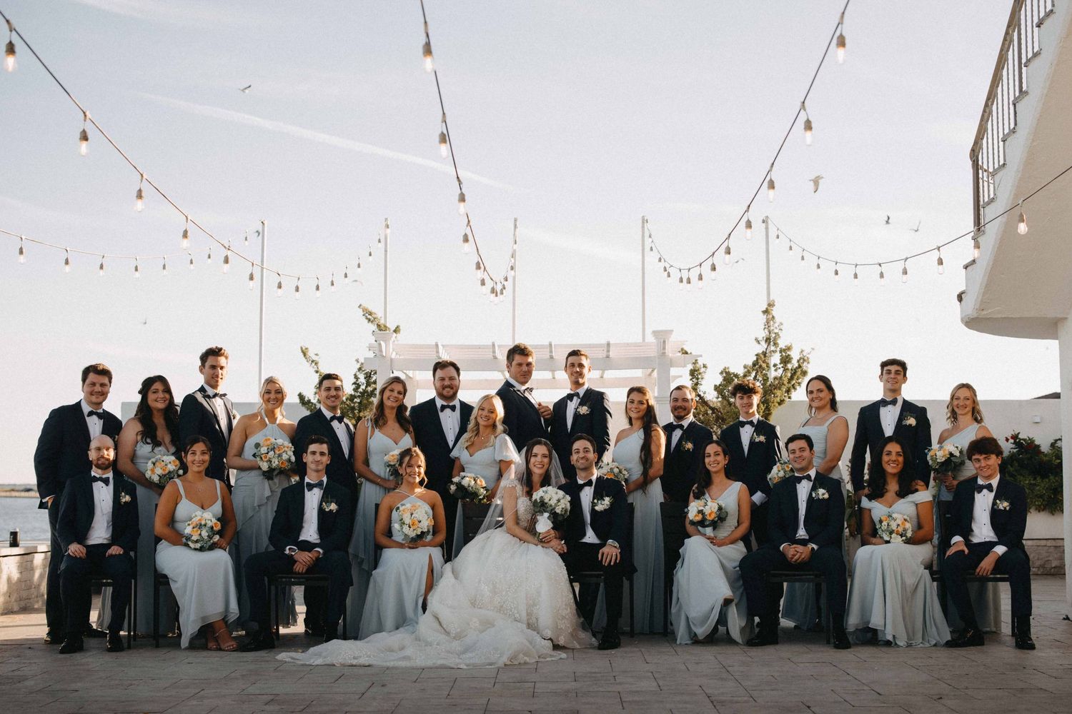 Large wedding party group photo on a rooftop terrace with string lights at dusk.