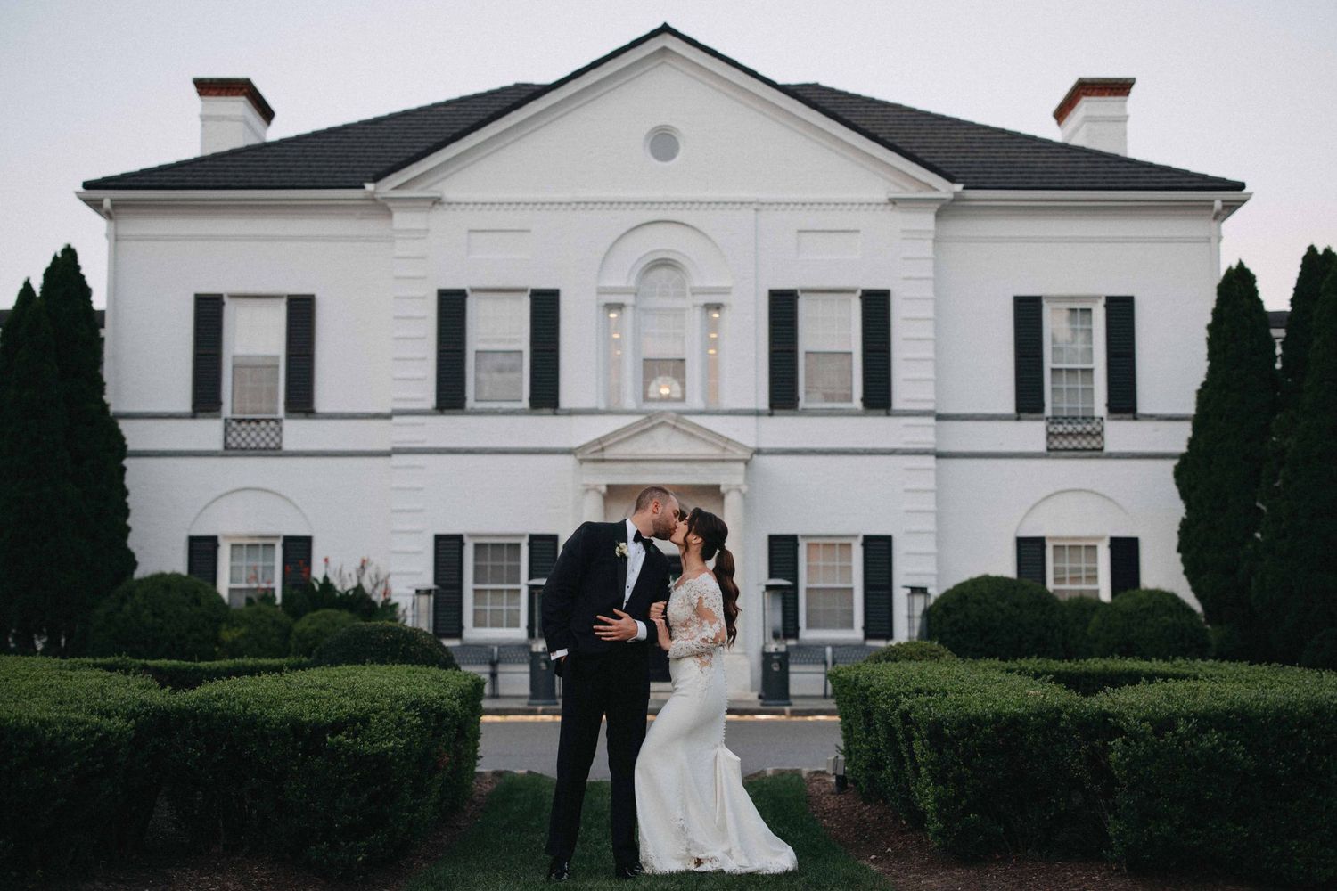 Bride and groom sharing a kiss in front of white colonial wedding venue at dusk.
