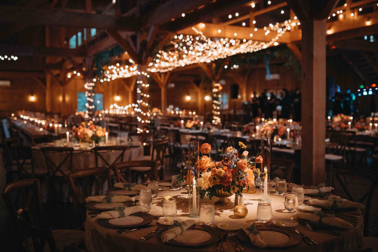 Warm rustic barn wedding reception with string lights, wooden beams, and autumn floral centerpieces.