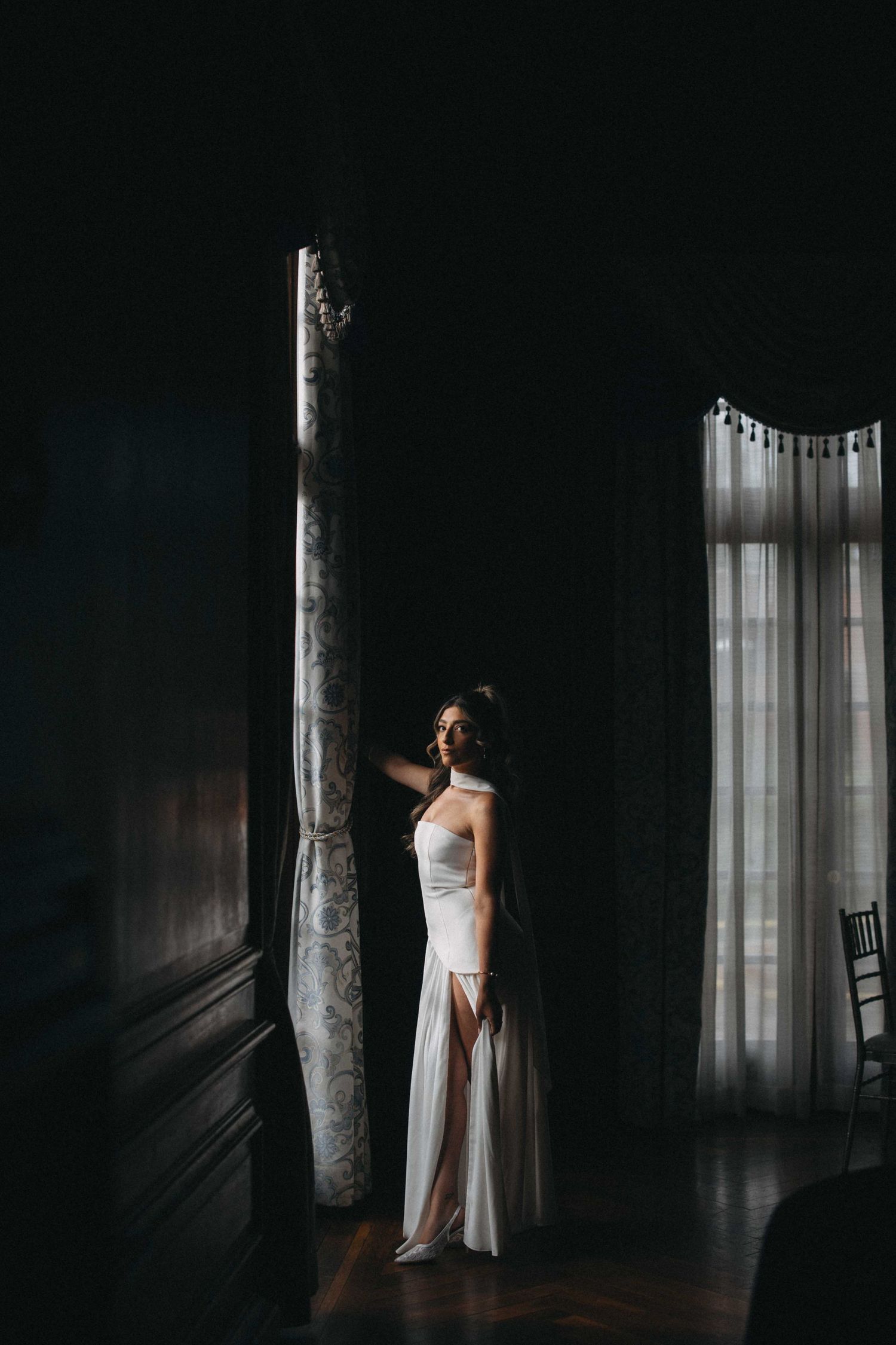 Bride in a white gown standing in dramatic shadow near a sunlit window indoors.