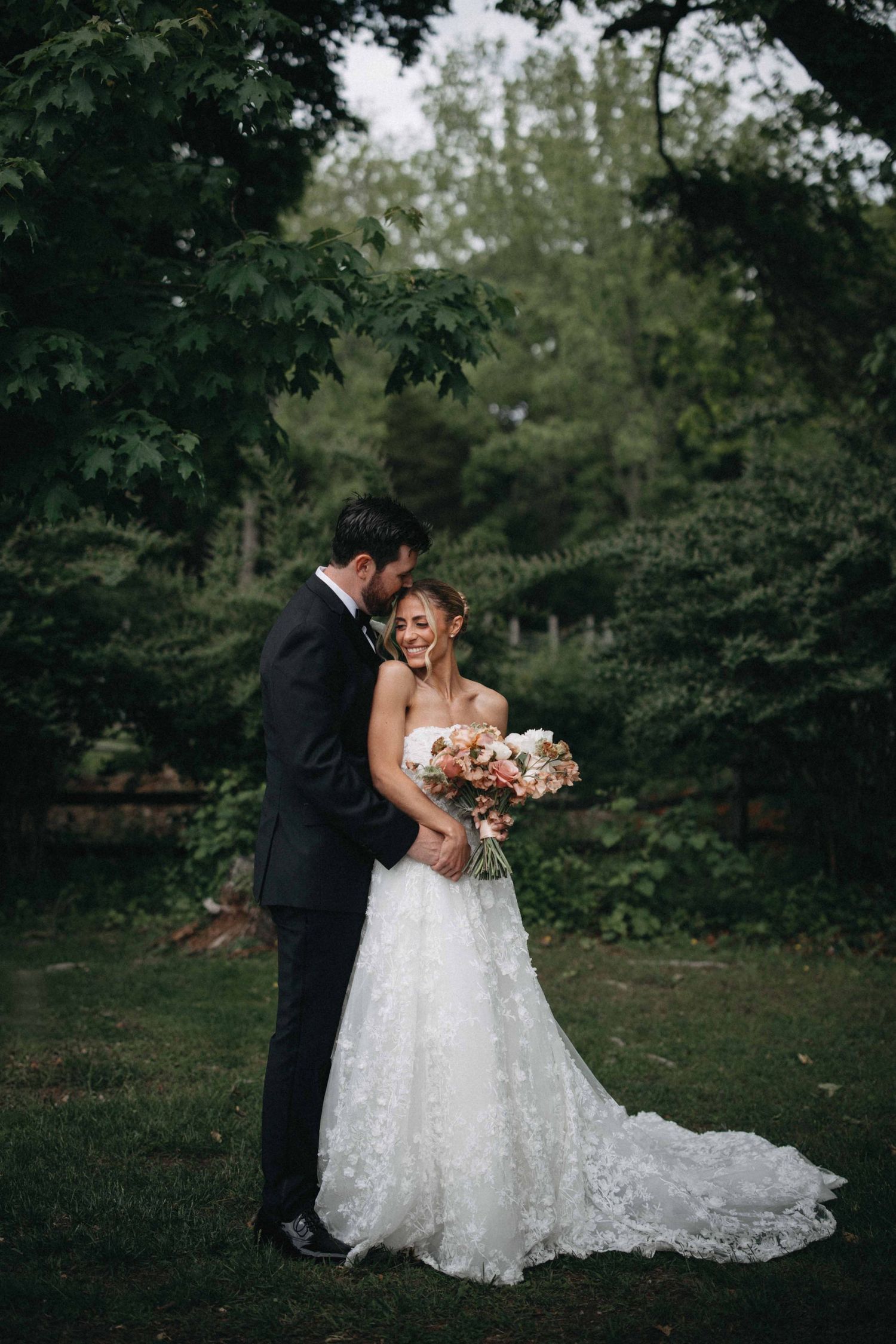 Bride in lace gown and groom in dark suit embracing in a lush green garden setting.