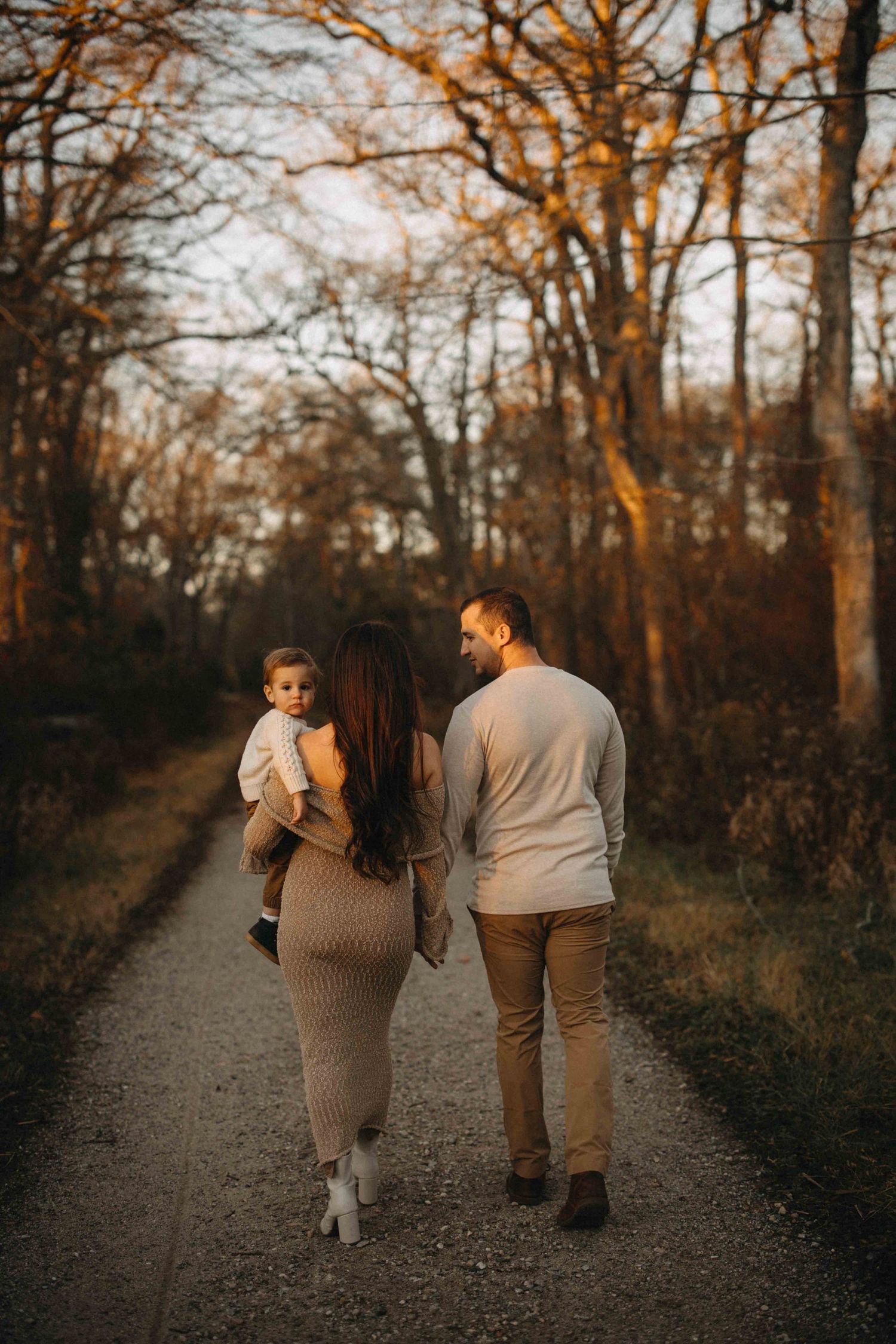 A young family walks along a scenic autumn path, mother holding baby while father looks on warmly at golden sunset.