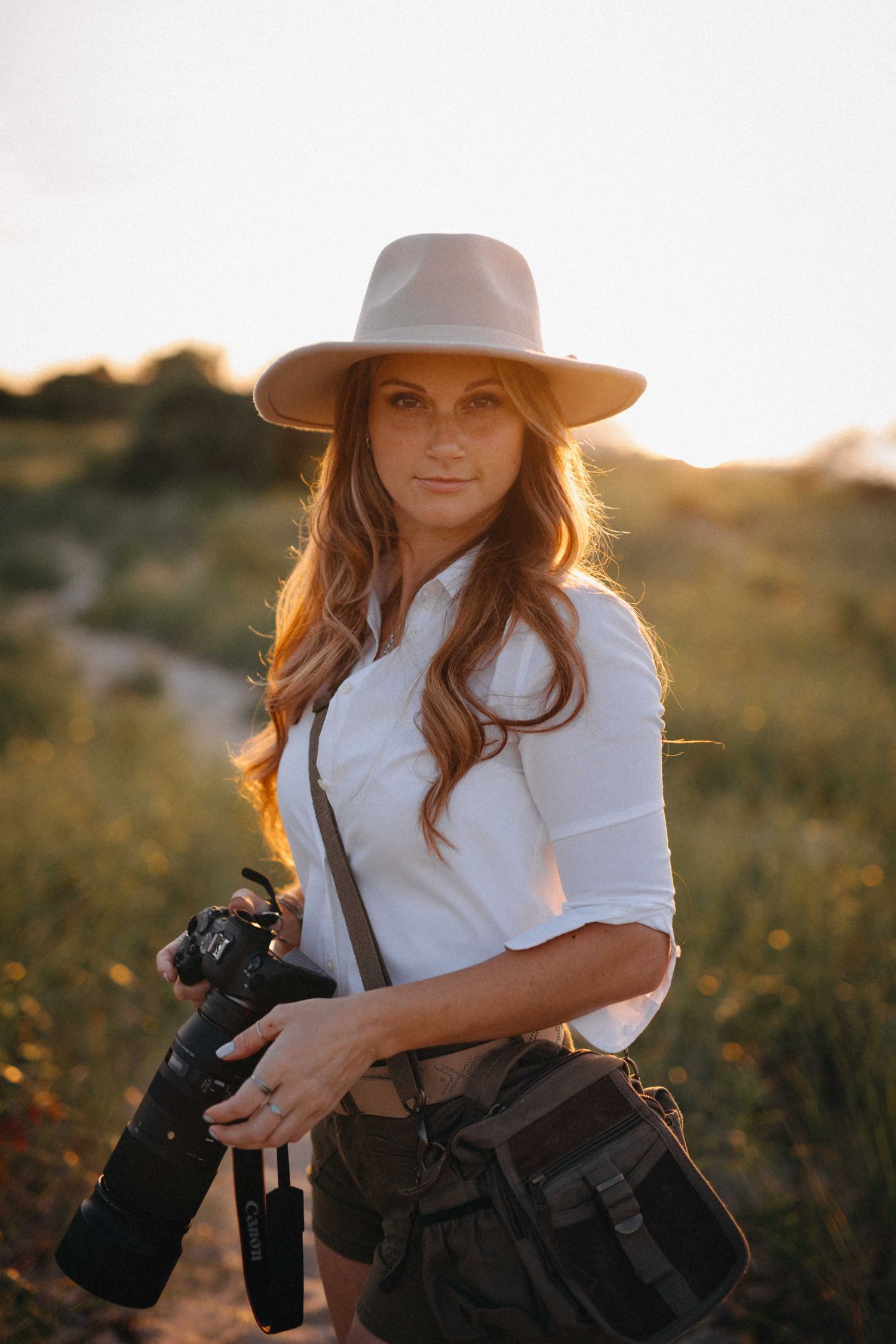 Female photographer in white shirt and wide-brim hat holding a camera in a golden sunset field.