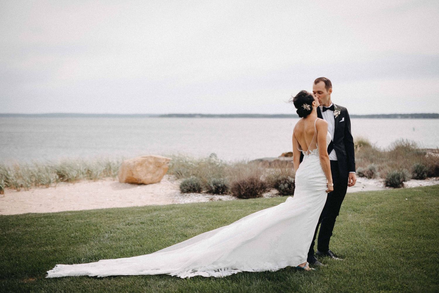 Bride and groom embracing on a manicured lawn with a sandy beach and calm ocean in the background.