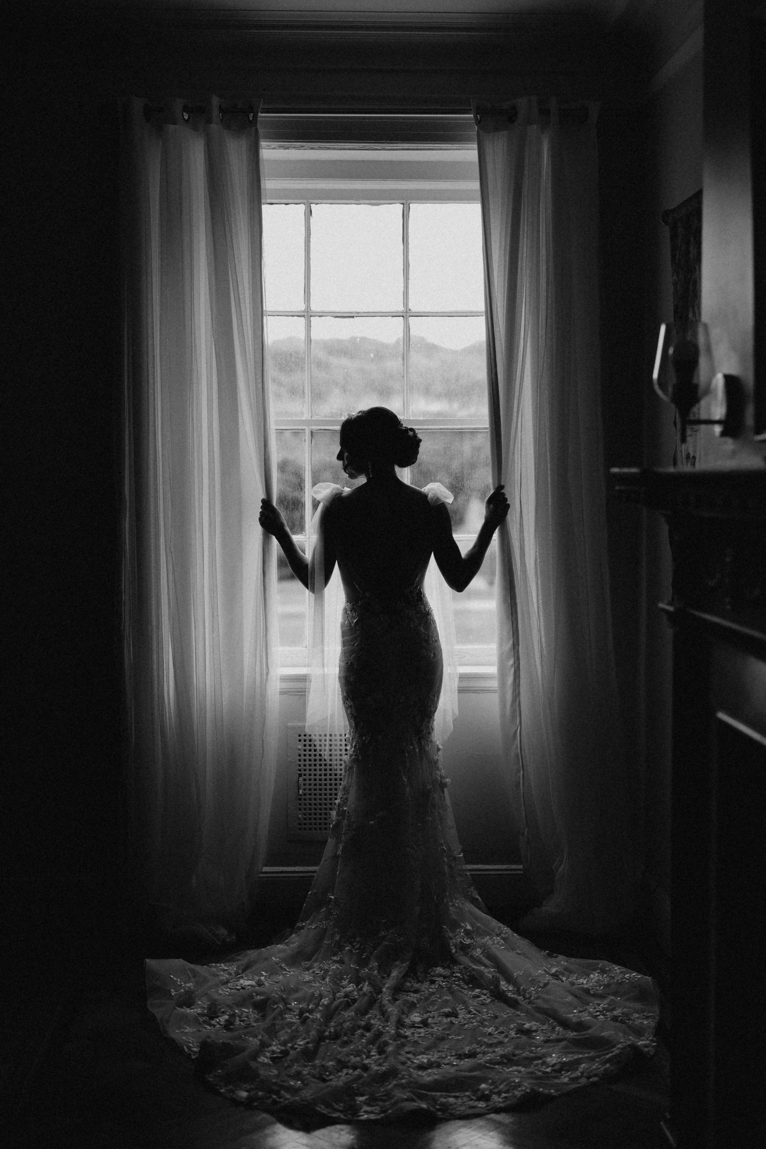 Silhouette of a bride in a lace mermaid gown standing before a large window in black and white.