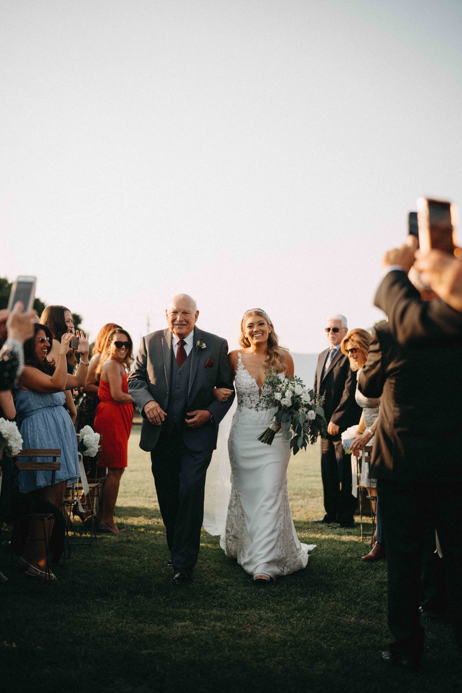 Bride in lace gown holding white bouquet walks down outdoor aisle with father at sunny wedding.