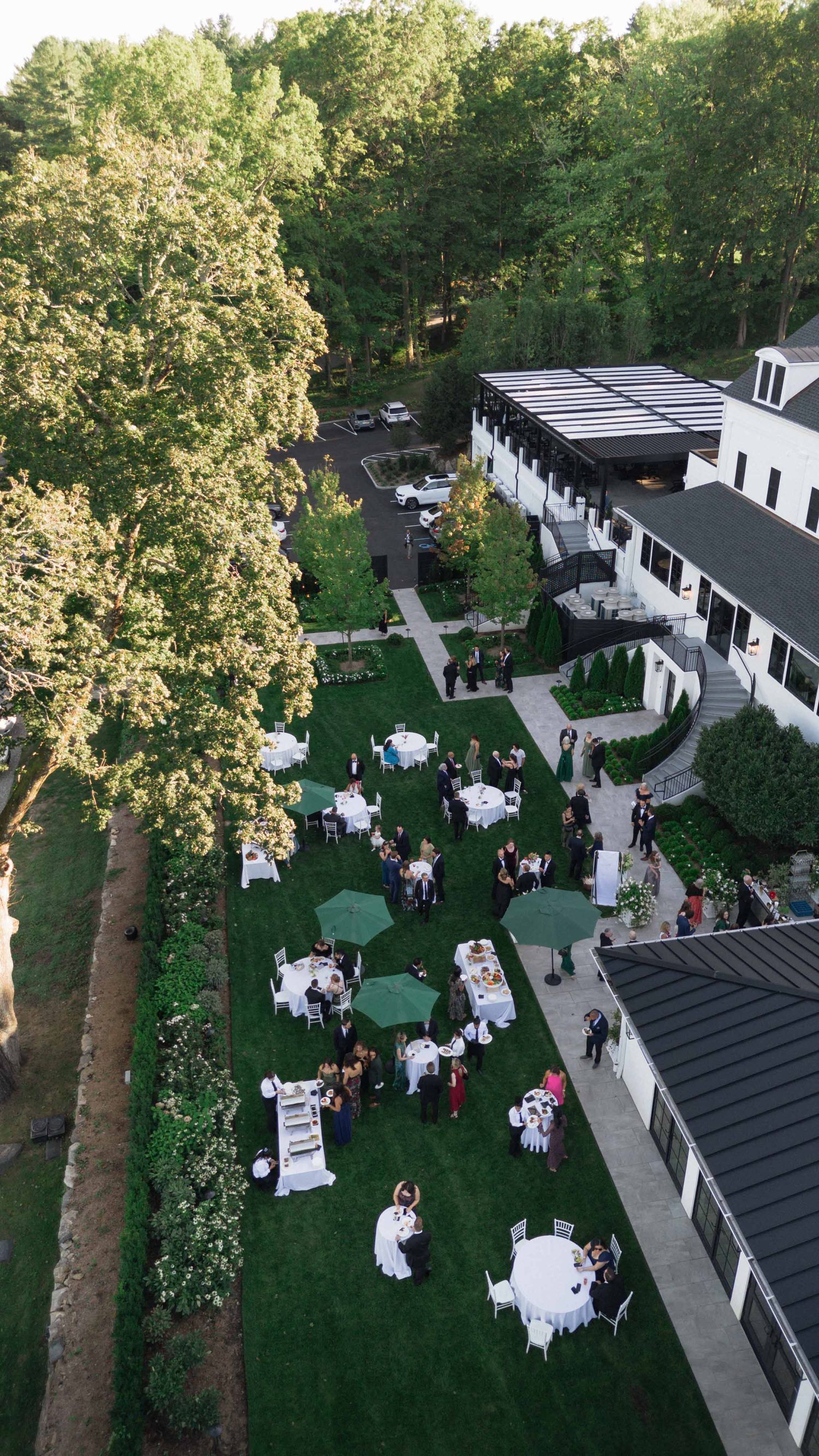 Aerial view of outdoor wedding reception with white tables and green umbrellas on manicured lawn beside white manor house.