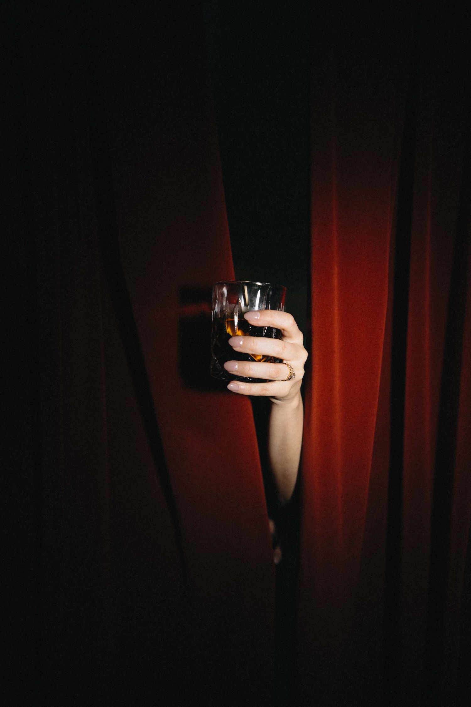 A hand emerges from red velvet curtains holding a glass of whiskey in dramatic dark lighting.