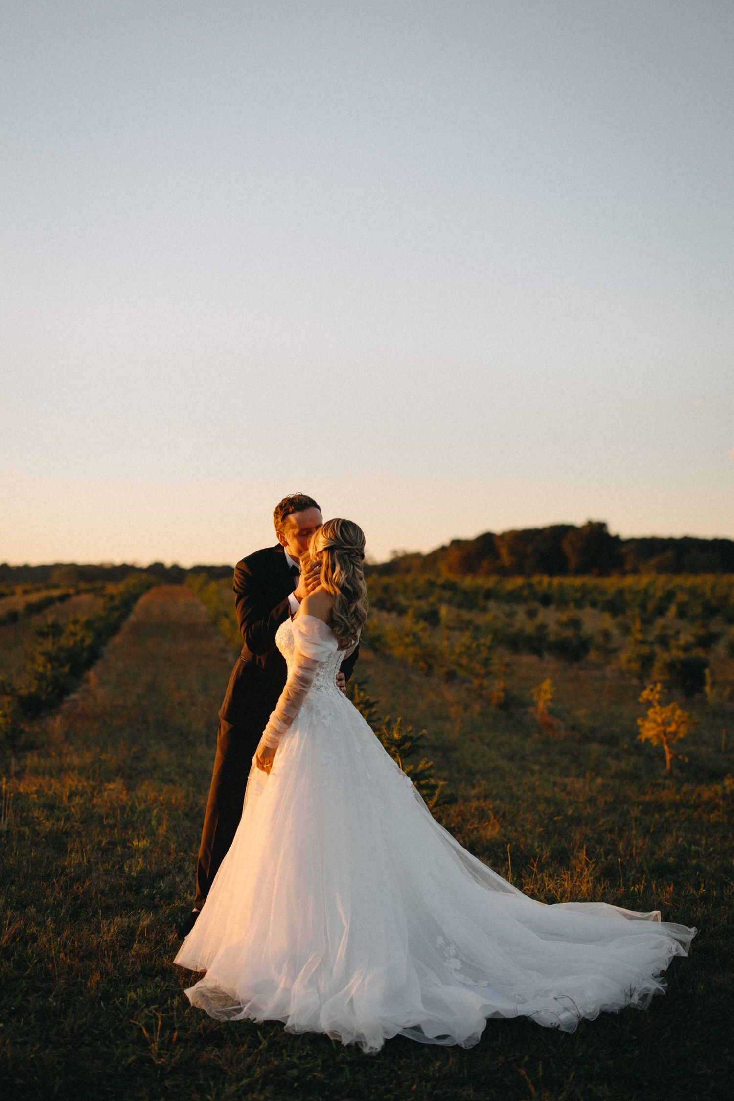Bride and groom silhouetted against glowing sunset in open field, bride's gown trailing behind.