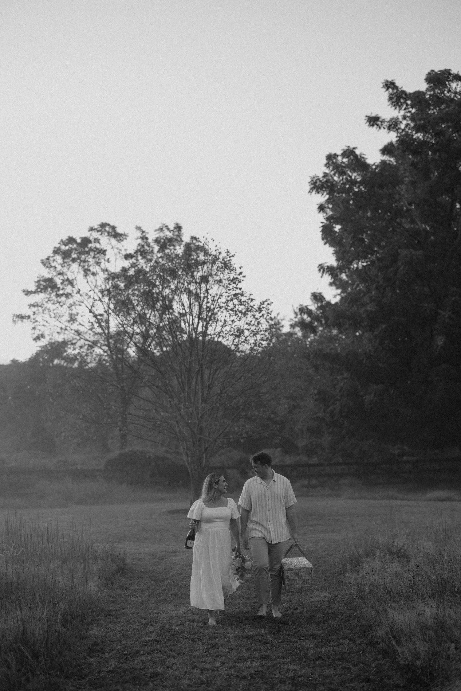 Black and white photo of a couple walking through a field at dusk, carrying a picnic basket among lush trees.