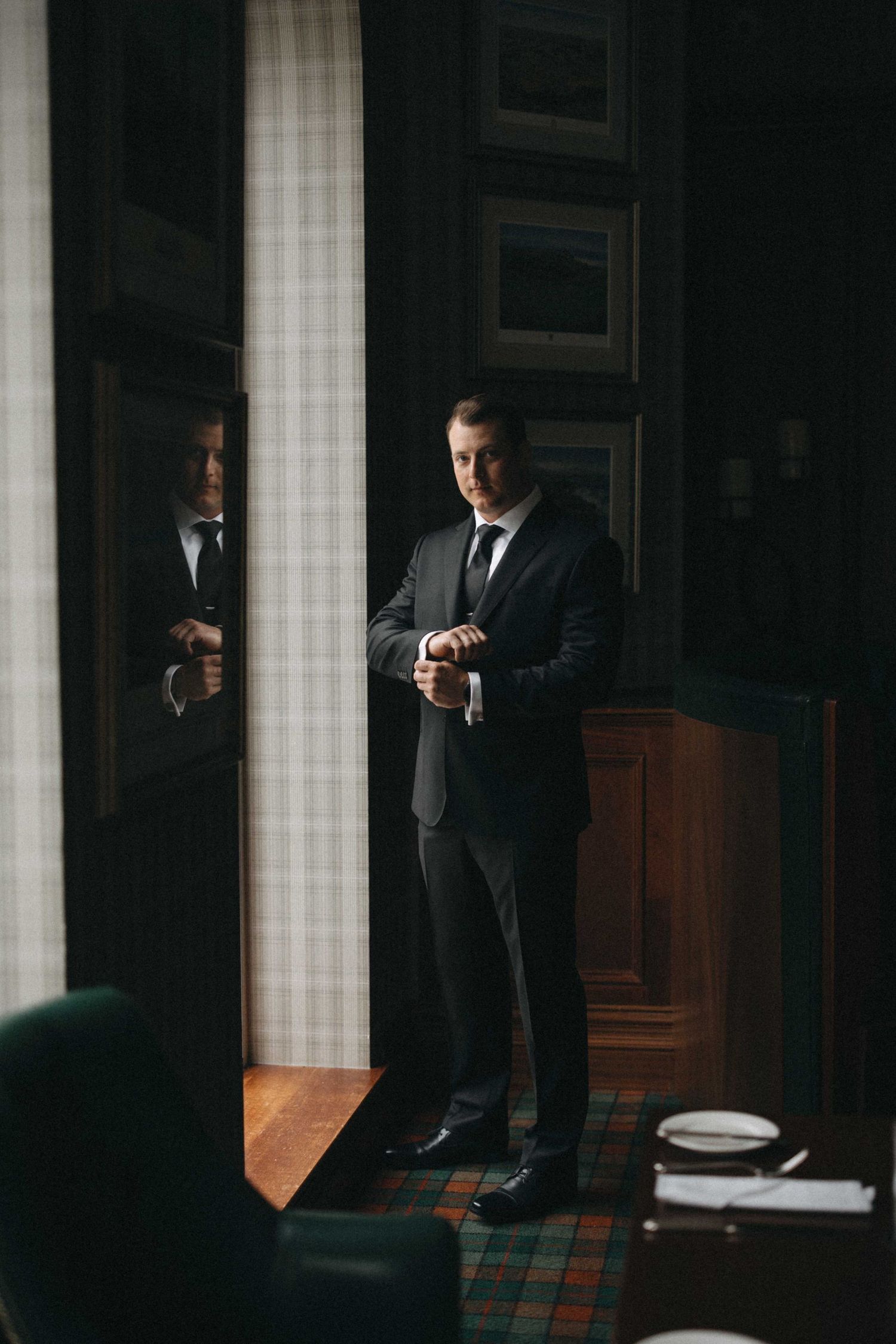 Dapper groom in a dark suit adjusts his cufflinks while standing near a mirror in an elegant wood-paneled room.