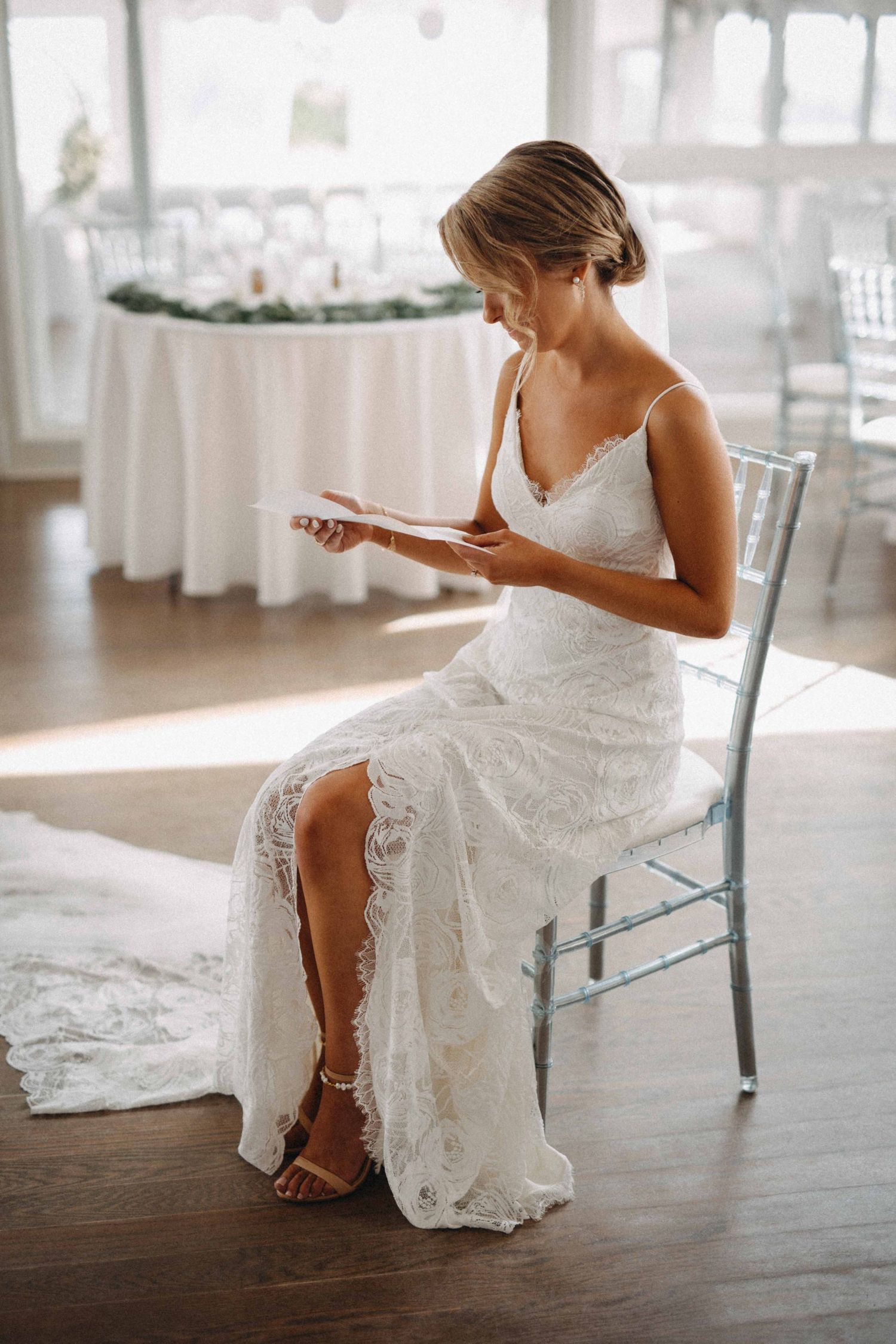 Bride in lace wedding gown reading vows while seated on chiavari chair in elegant reception venue.