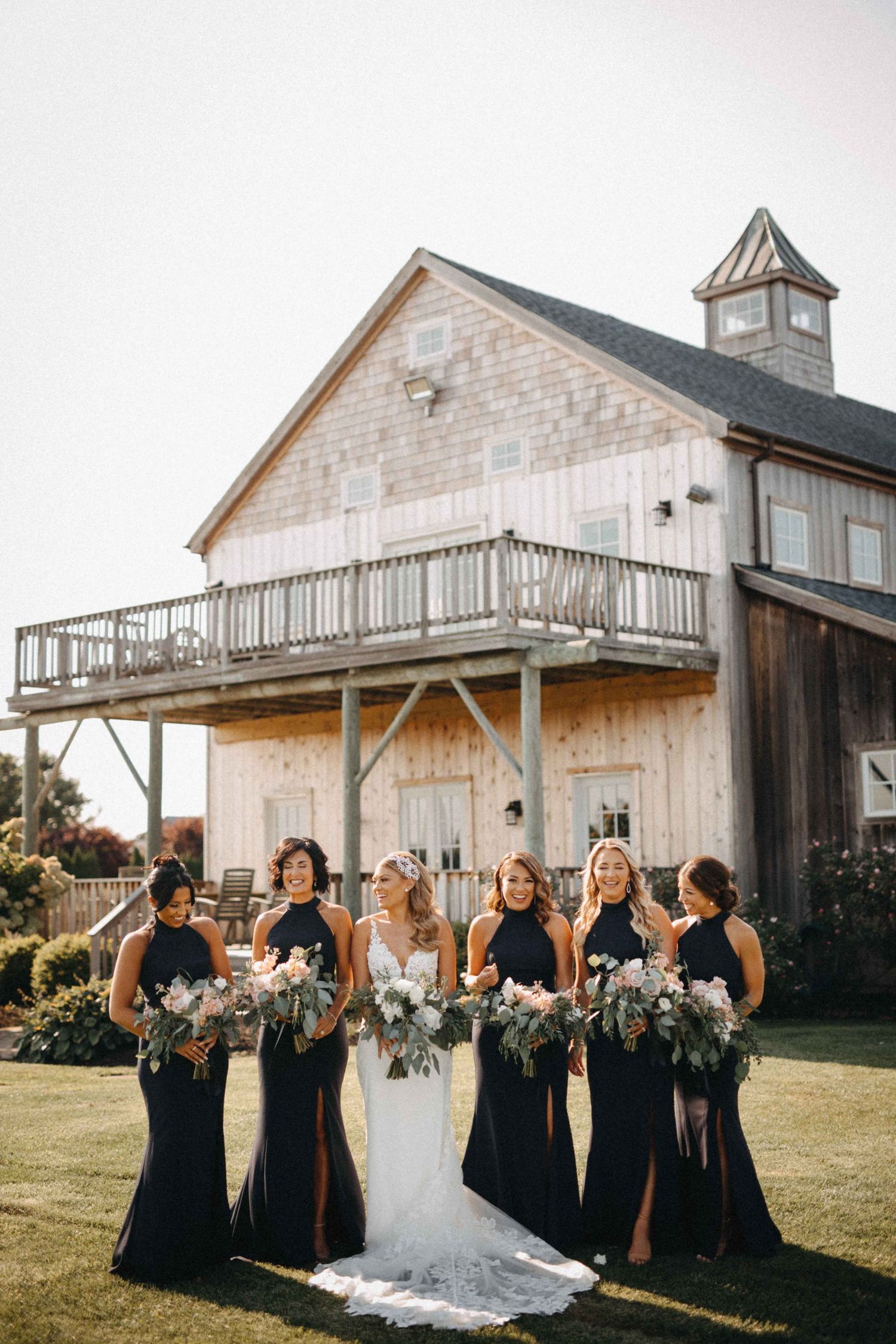 Bridesmaids in navy halter gowns holding bouquets in front of rustic barn venue.