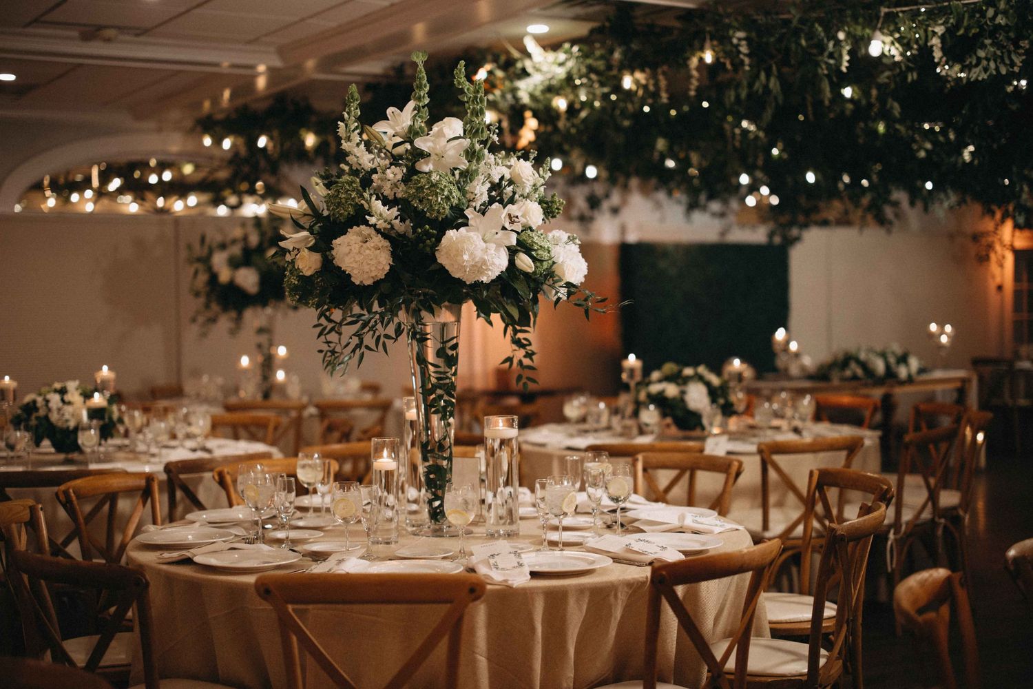Elegant wedding reception hall with tall floral centerpieces of white roses and greenery, warm string lights overhead.