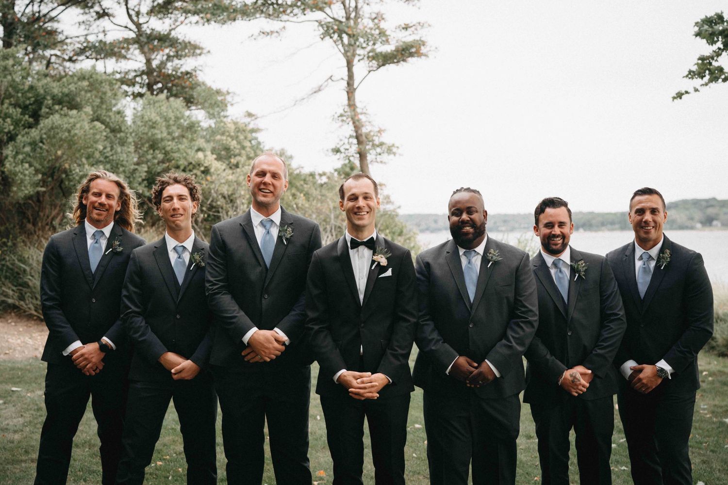 Groom and groomsmen in dark suits with light blue ties posing outdoors near waterfront.