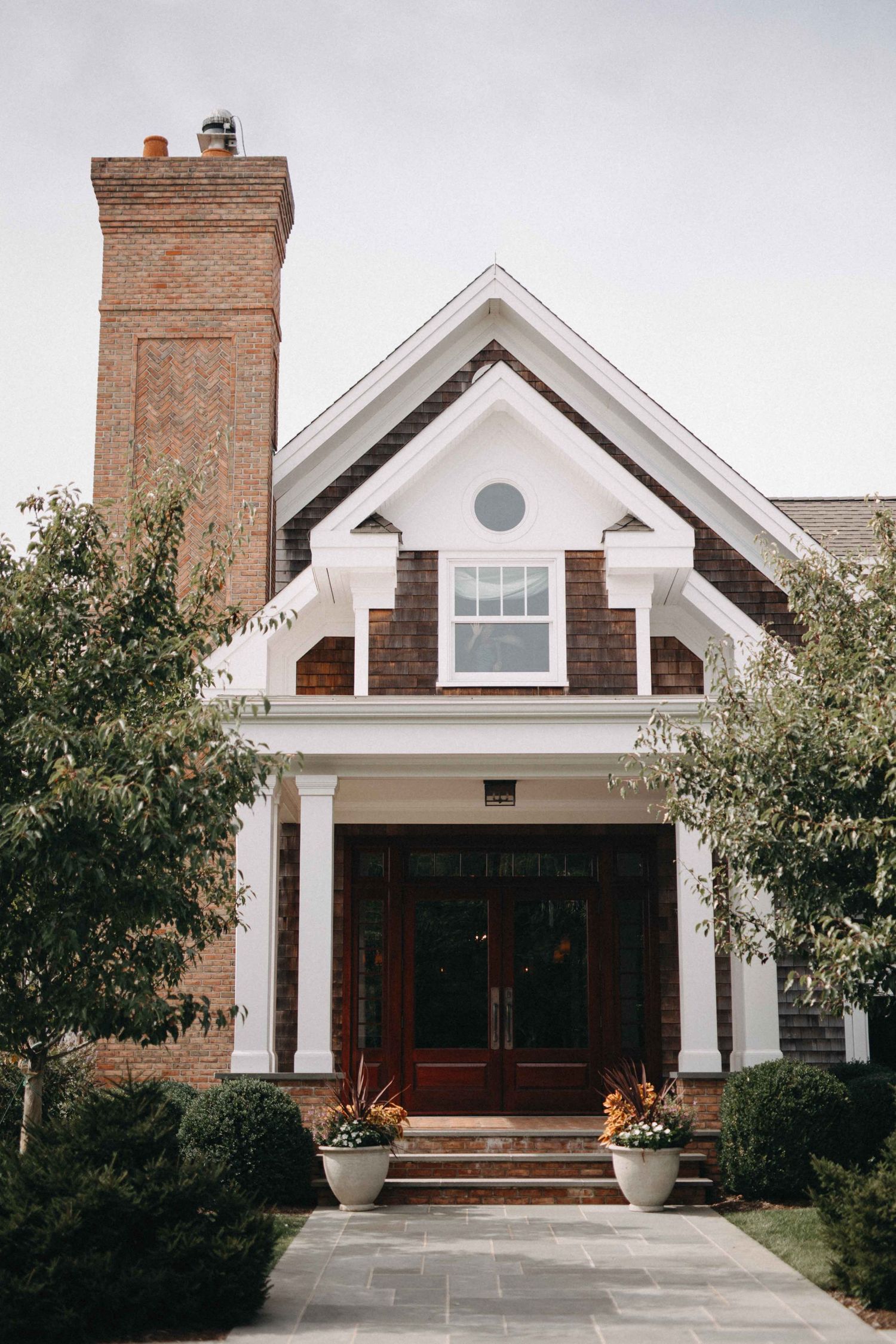 Elegant craftsman-style home exterior with brick chimney, round window, white columns, and red double doors.