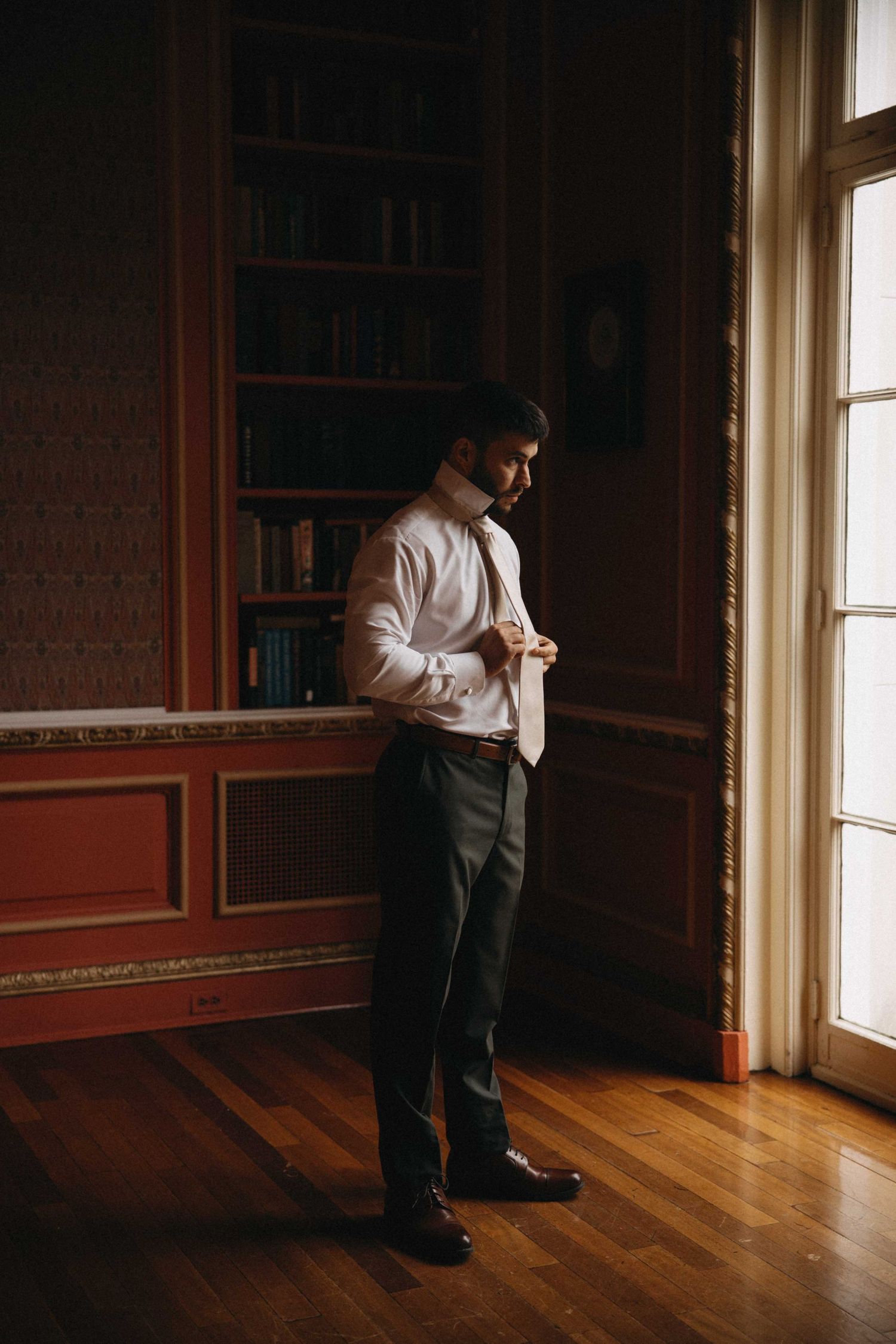 Groom adjusts his tie in a dark wood-paneled library room with bookshelves while preparing for his wedding day.
