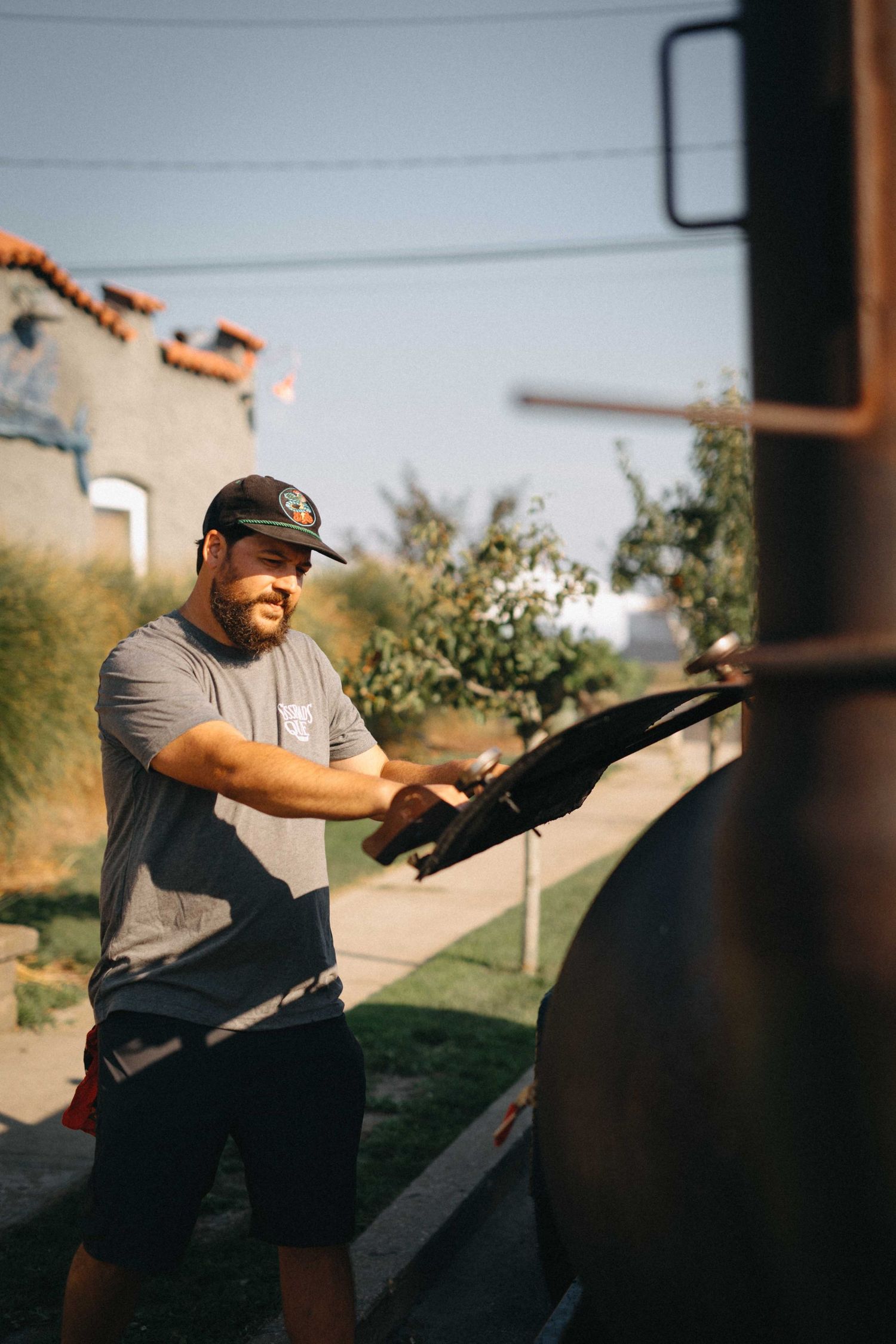 Bearded man in gray shirt and cap opening a large outdoor smoker grill on a sunny suburban street.