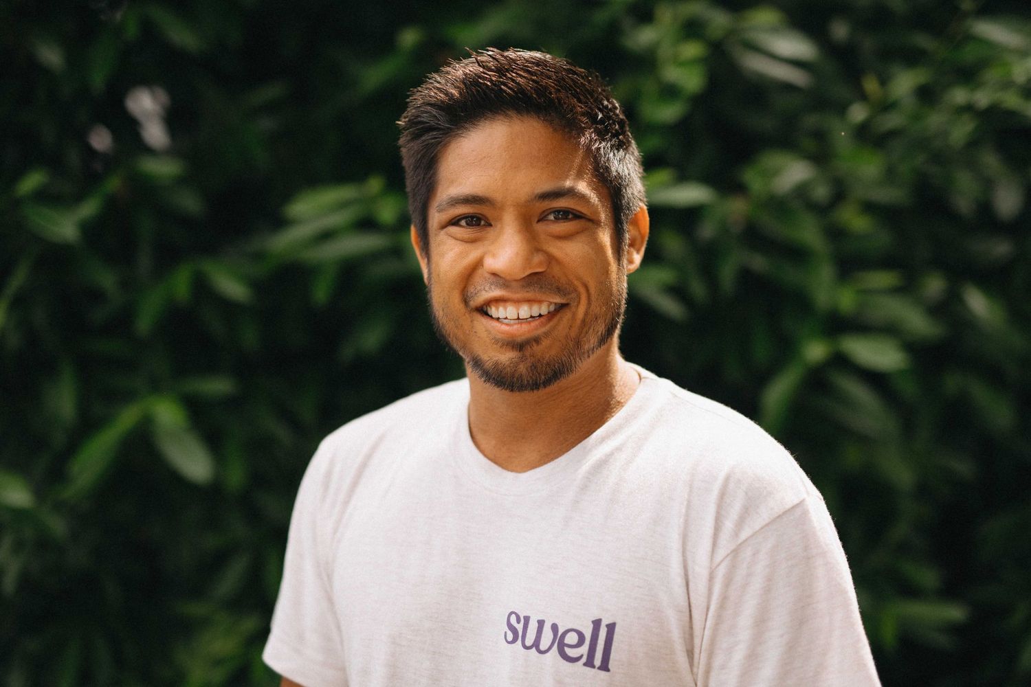 Smiling man wearing white Swell t-shirt outdoors with lush green foliage background.