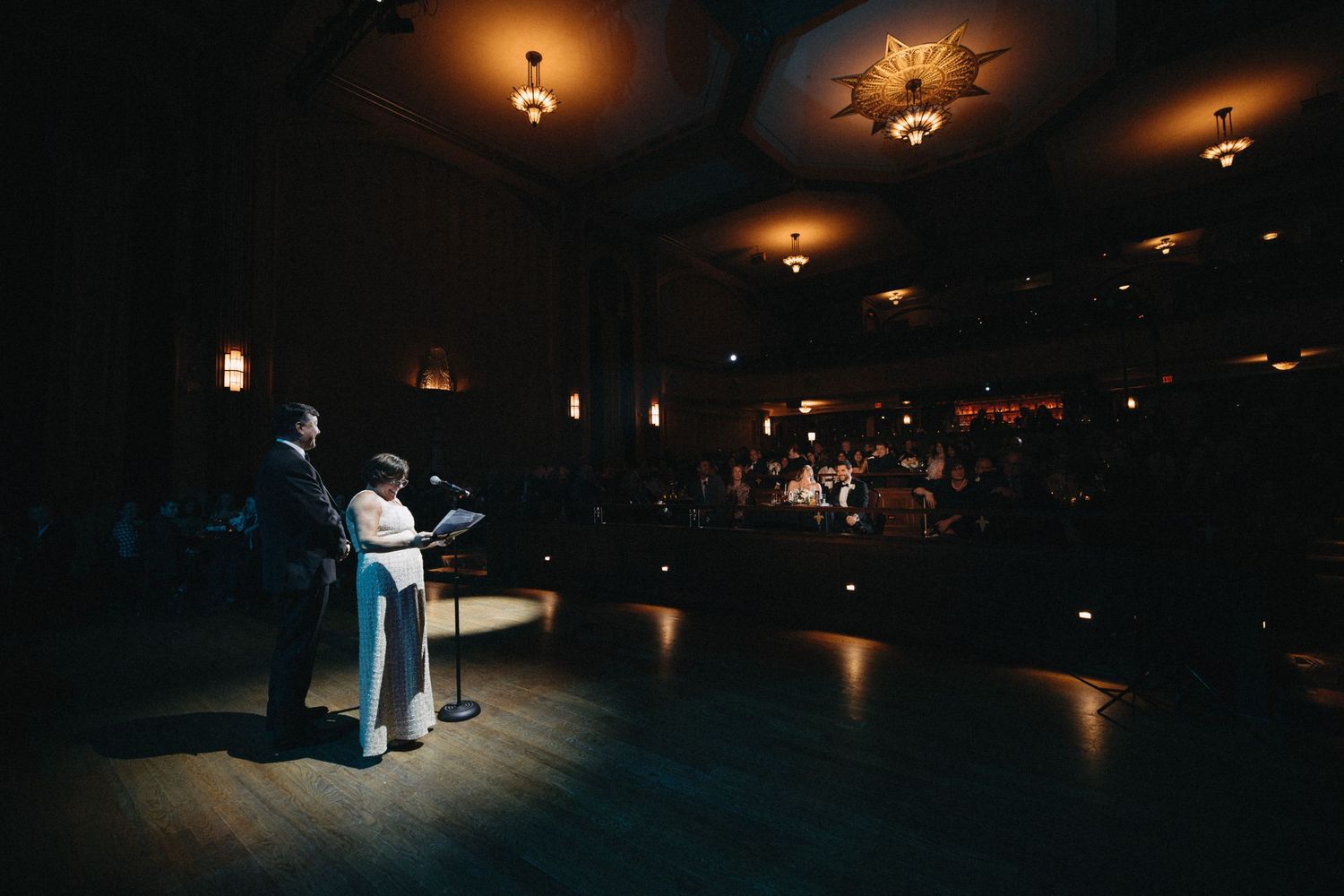 Couple speaks at podium on stage before crowded wedding reception in grand historic theater venue.