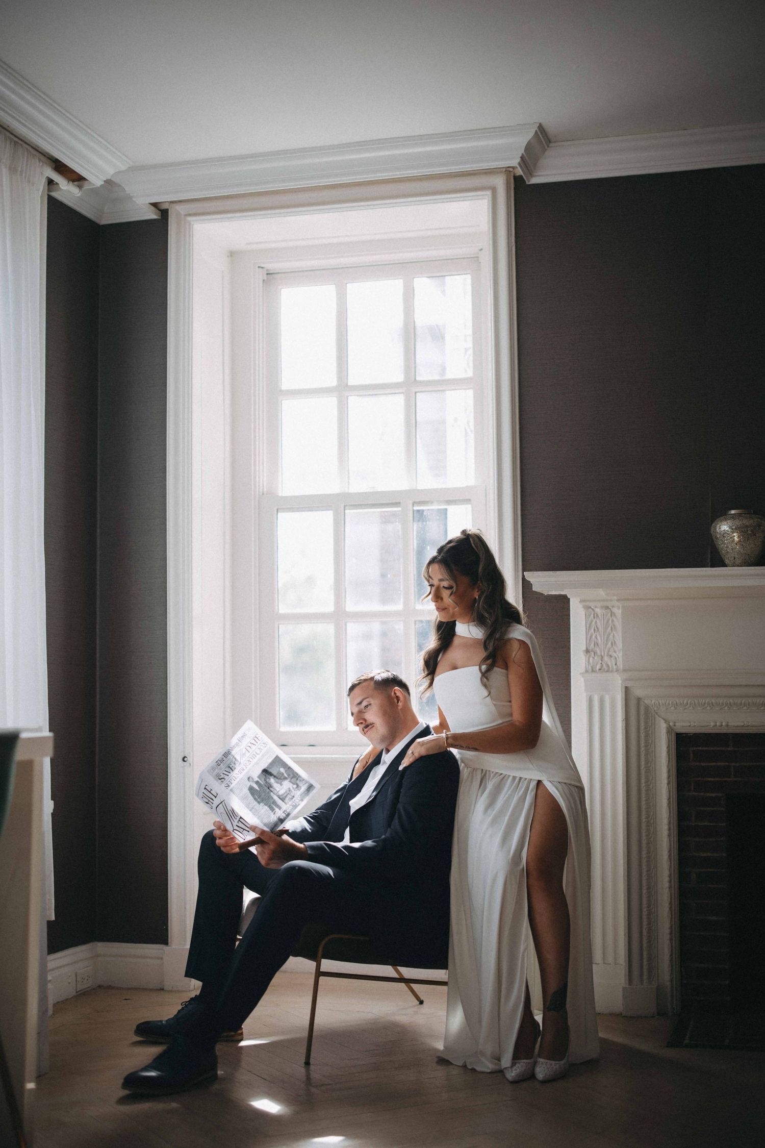 Groom reads newspaper in chair while bride in white gown stands by window.