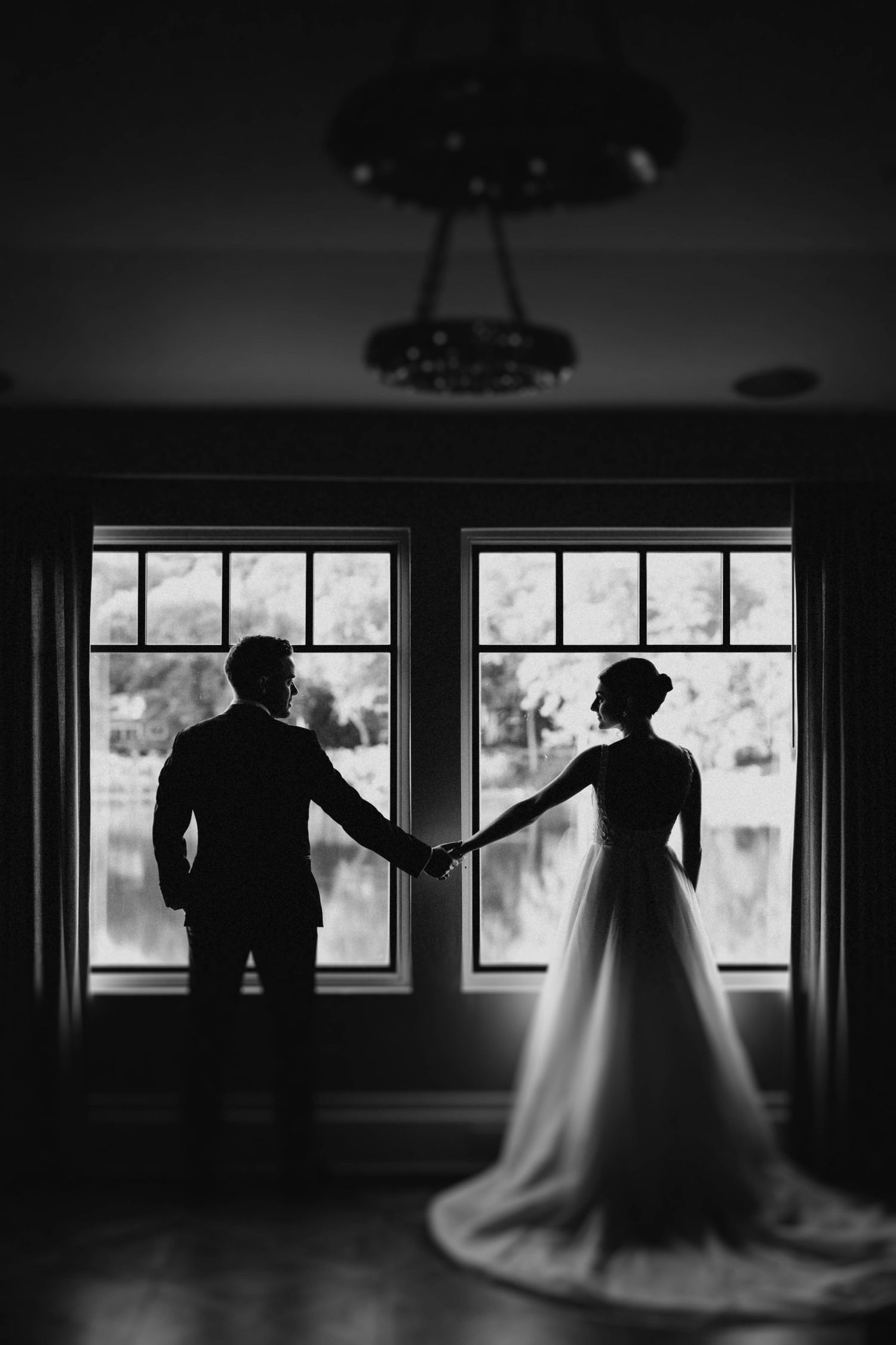 Silhouette of bride and groom holding hands in front of large windows in a black and white wedding portrait.