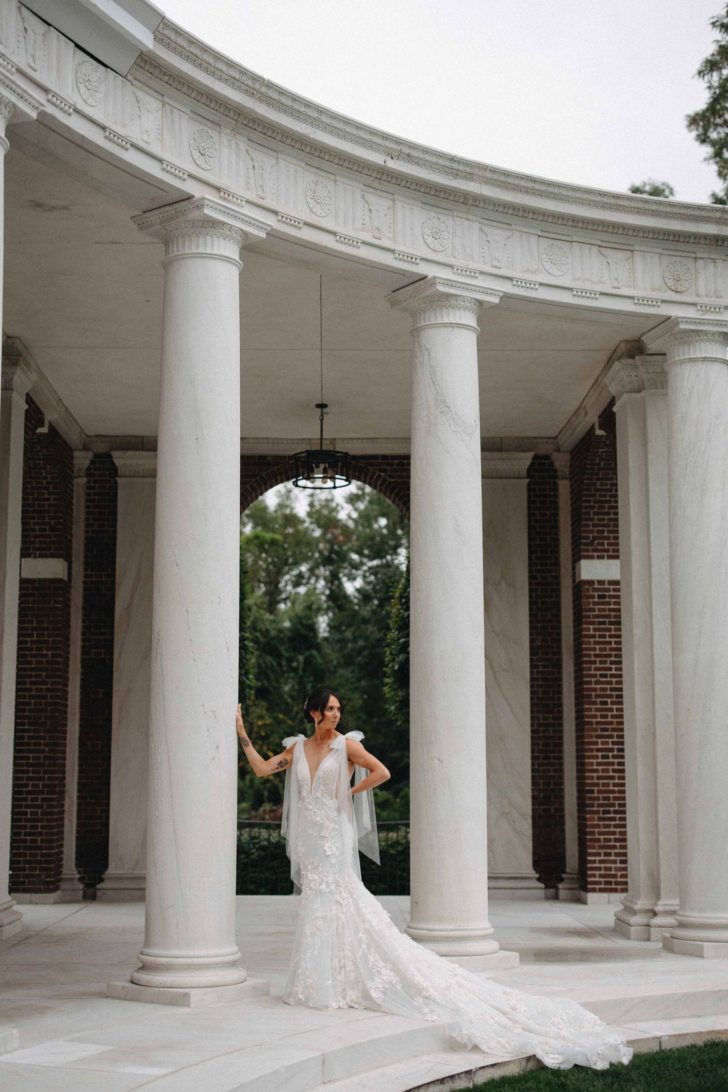 Bride in lace mermaid gown with veil poses dramatically among white columns of a grand rotunda.