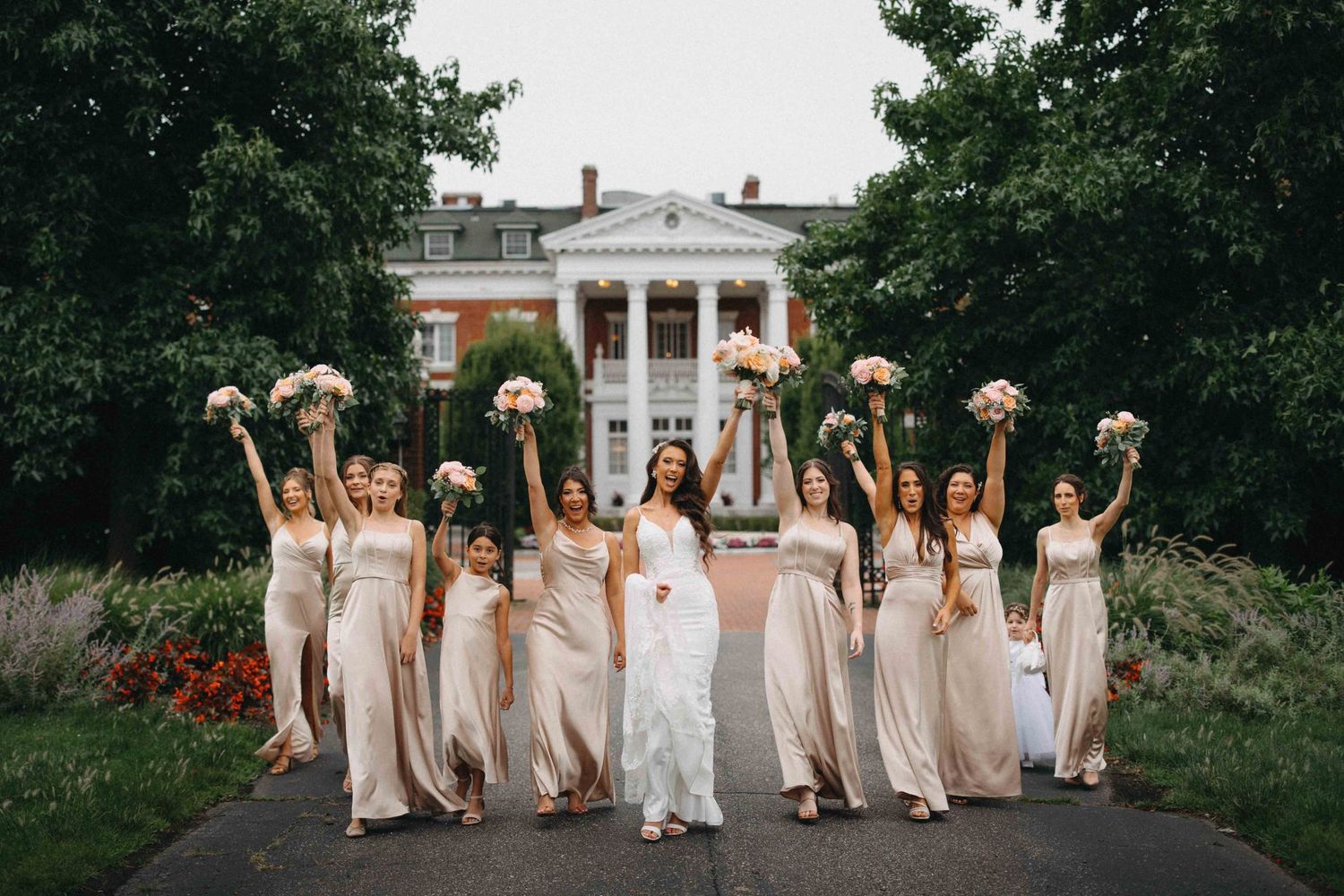 Bride and bridesmaids in champagne gowns raising bouquets in front of elegant white mansion.