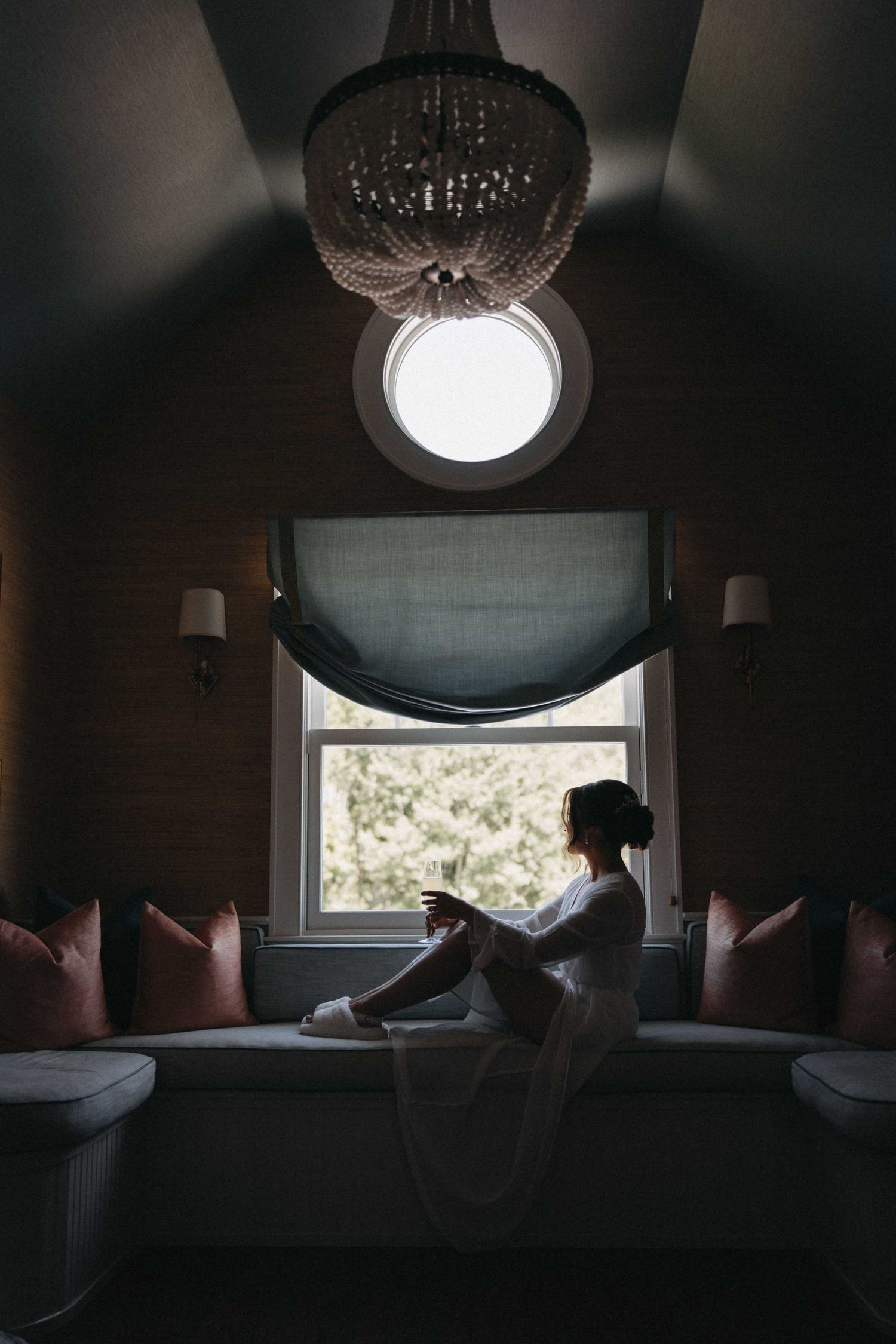 Bride in white robe sits peacefully by window in elegant room with crystal chandelier.