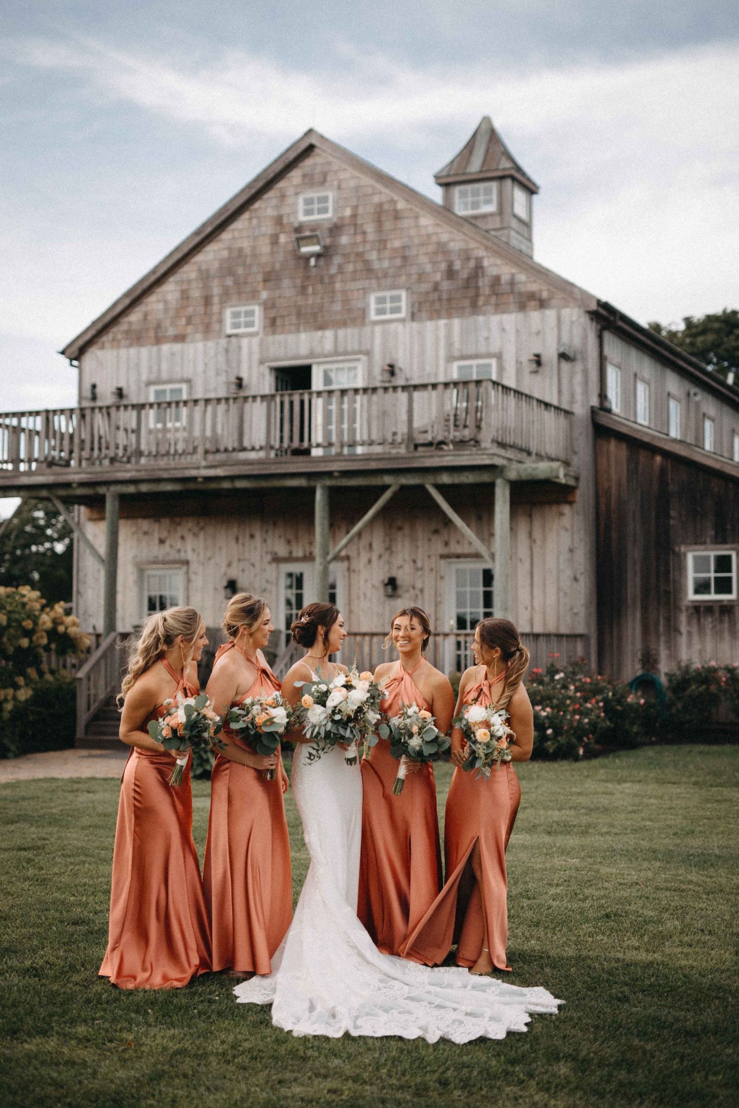 Bridesmaids in terracotta gowns holding bouquets stand with bride in white dress before rustic barn wedding venue.
