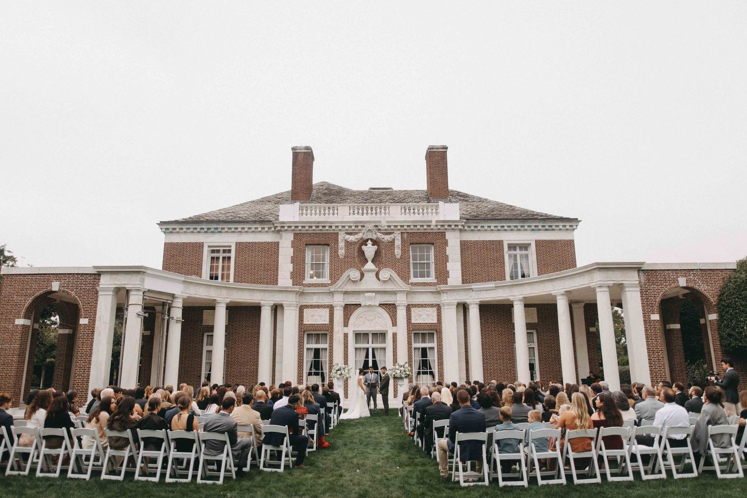 Wedding ceremony on manicured lawn in front of a grand brick colonial mansion with white columns and seated guests.