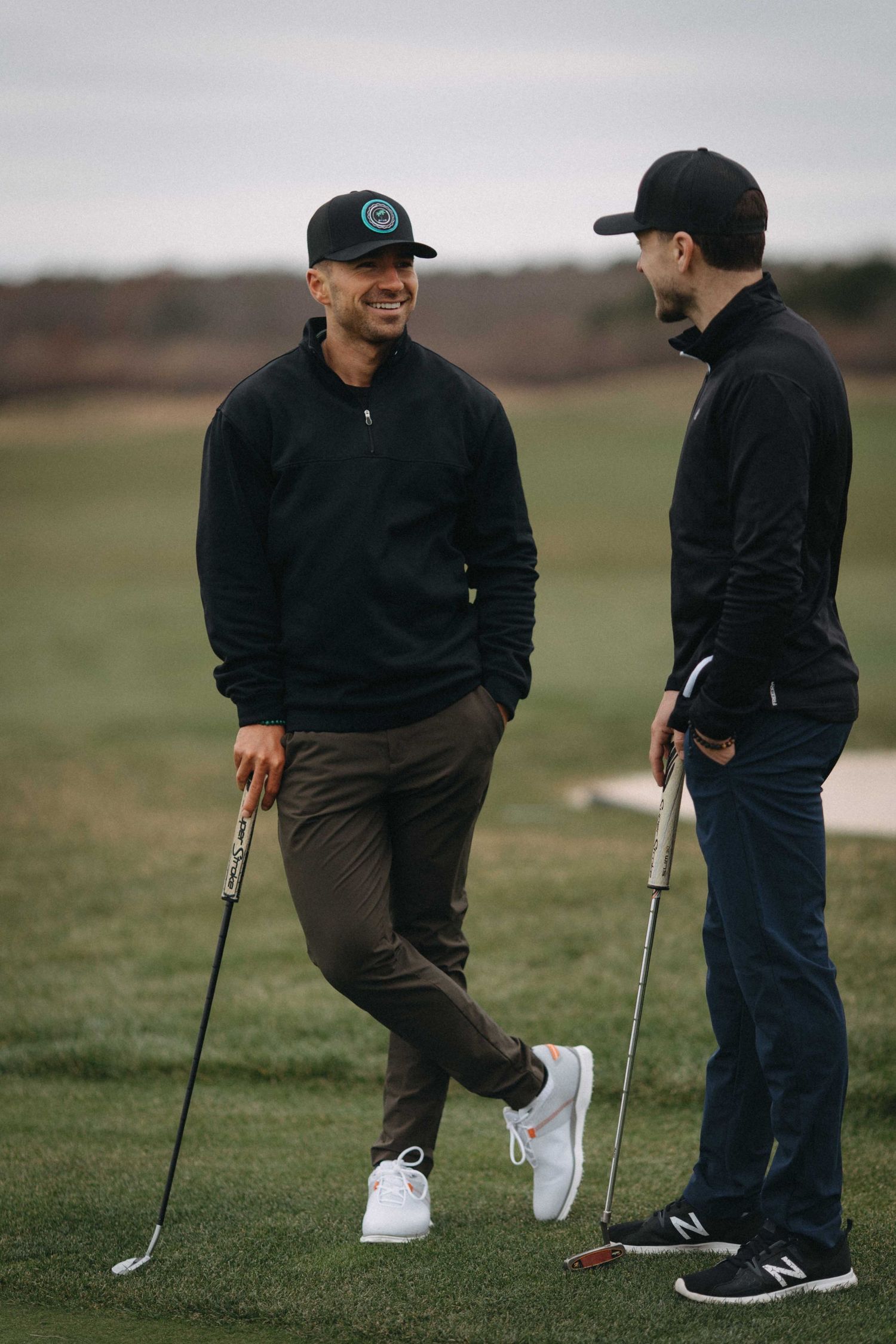 Two men in black athletic wear holding golf clubs and chatting on a golf course on a cloudy day.