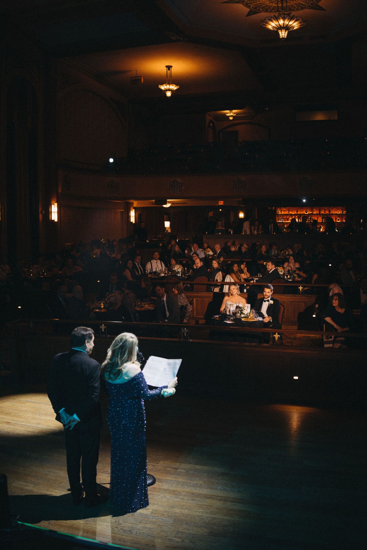 Wedding officiant speaks on stage before seated guests in grand historic theater venue.