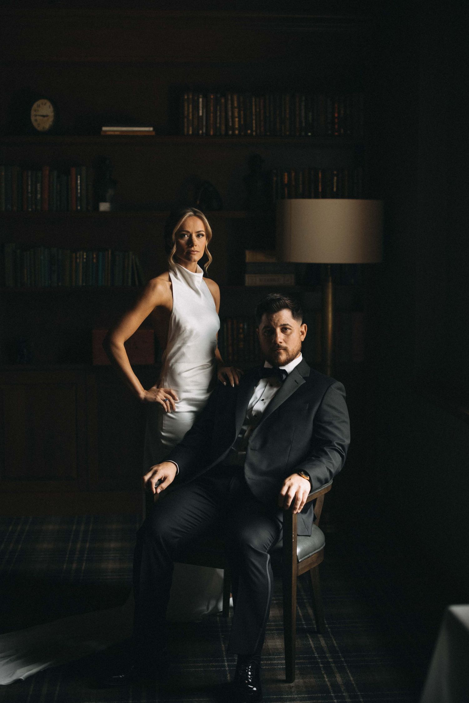 Elegant couple in formal attire posing in a dimly lit library setting for wedding portraits.
