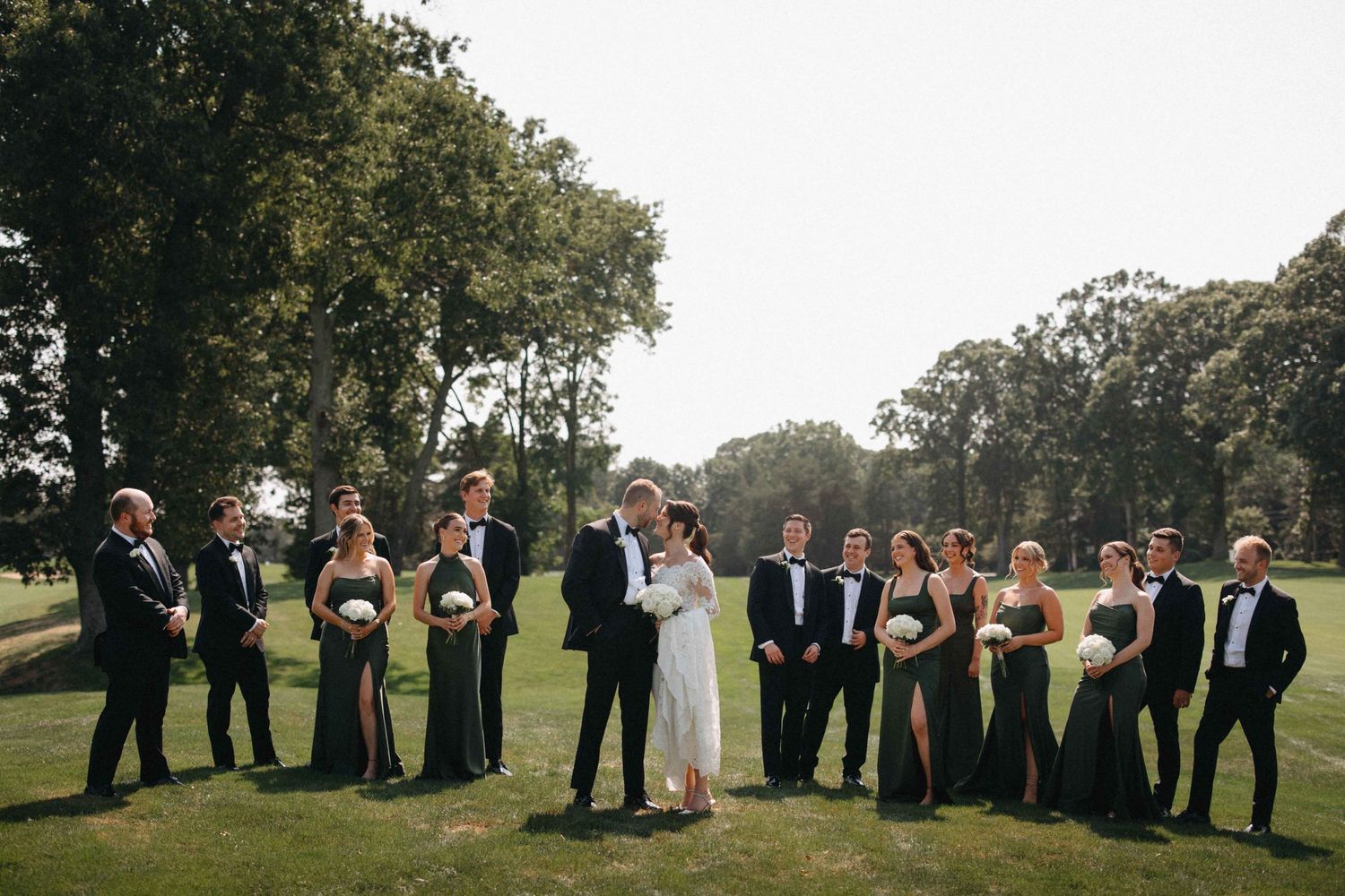 Wedding party in black tuxedos and dark green gowns gather on sunny golf course as couple kisses.