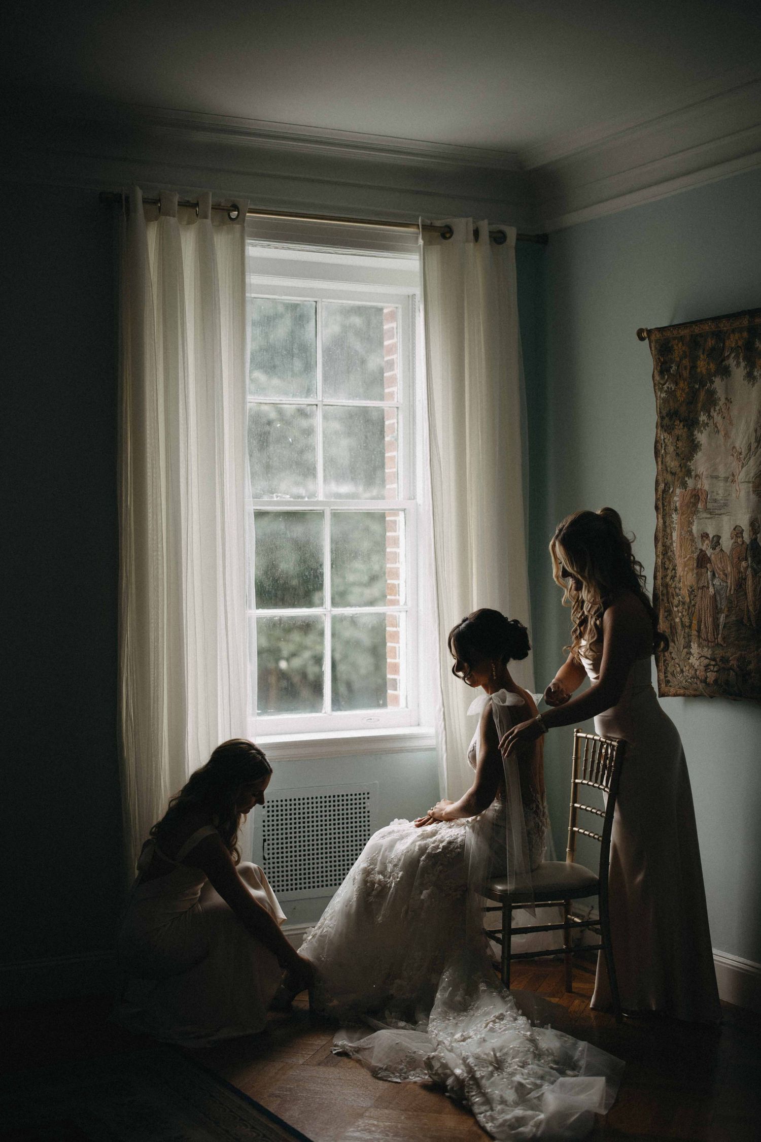 Bridesmaids help bride in lace gown prepare by a sunlit window in a softly lit vintage room.