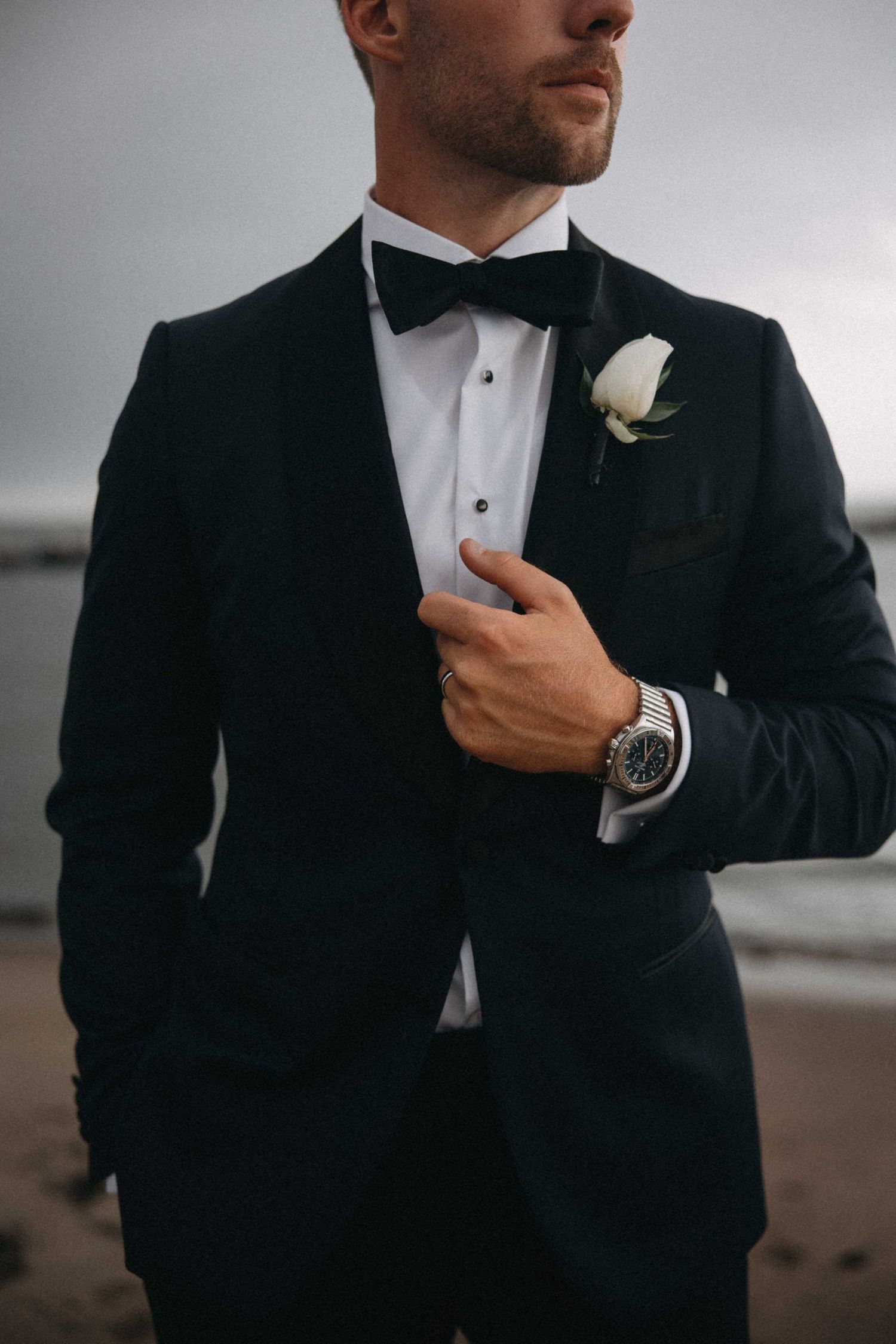 Dapper groom in black tuxedo with bow tie, white boutonniere, and luxury watch at beach wedding.