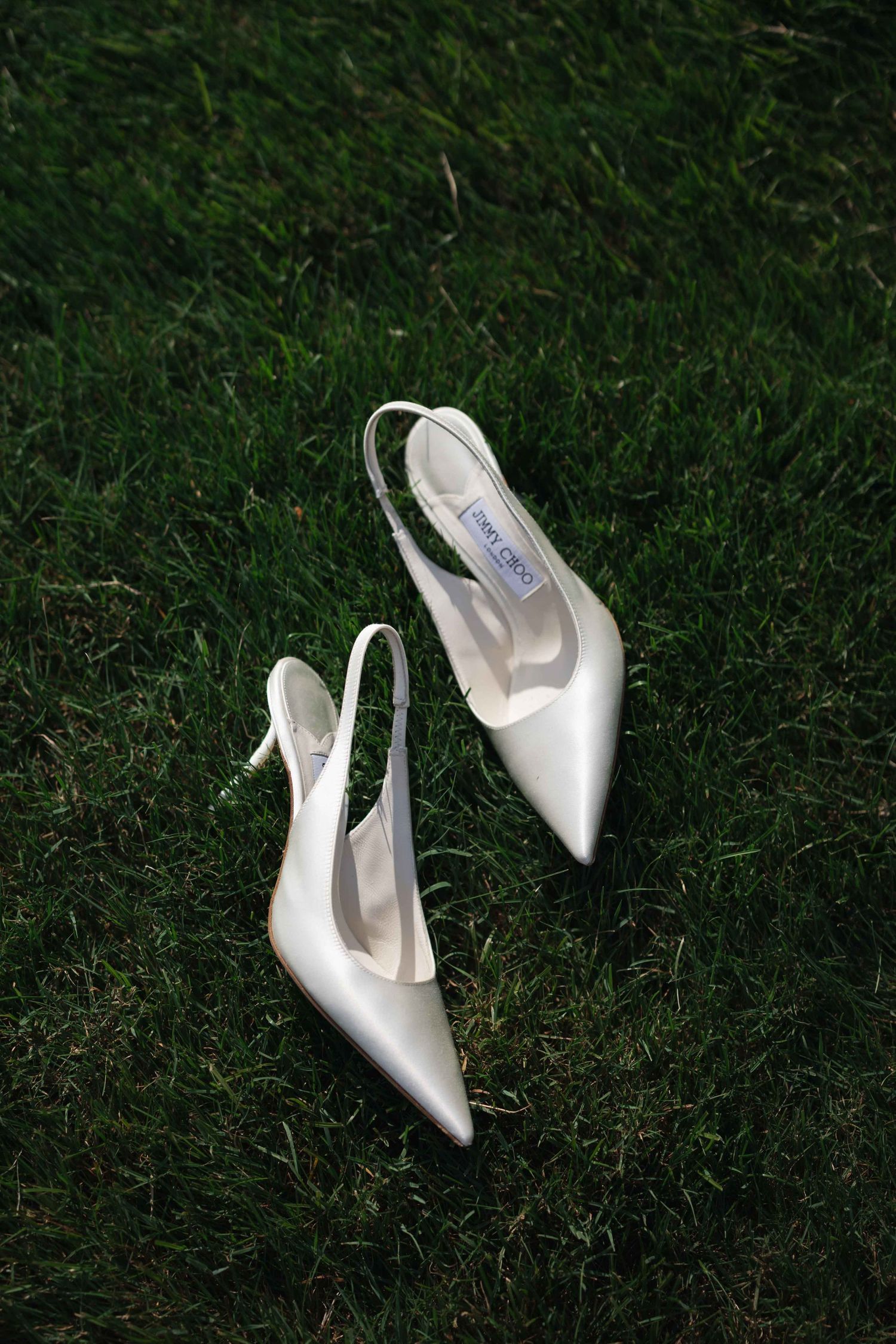 White Jimmy Choo slingback pointed-toe heels resting on lush green grass, perfect bridal wedding shoes.