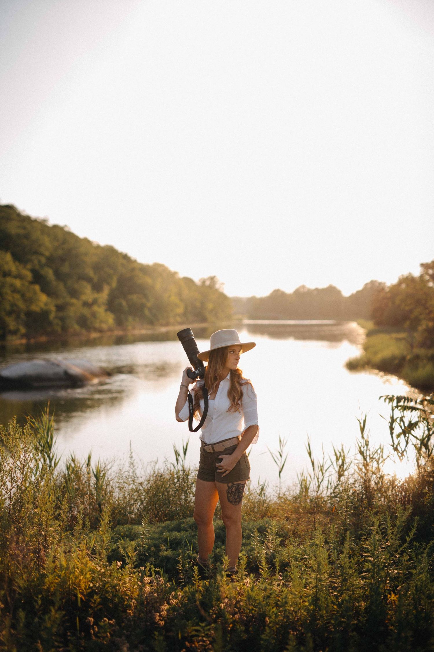 Woman in white hat and shirt holding camera near scenic river surrounded by lush trees.