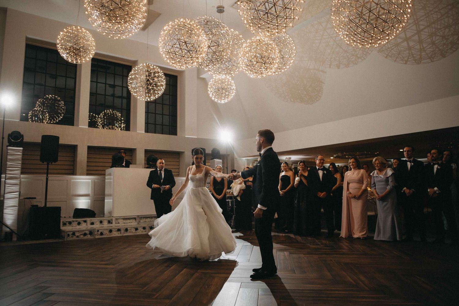 Bride and groom sharing first dance under glowing spherical chandeliers with guests watching.