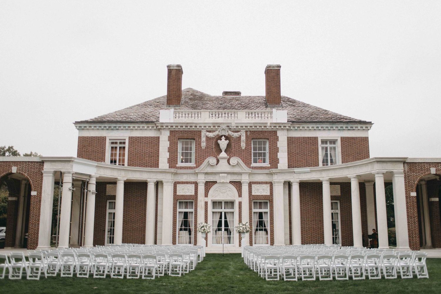 Grand brick colonial mansion with white columns serves as a stunning outdoor wedding ceremony venue with rows of white chairs.