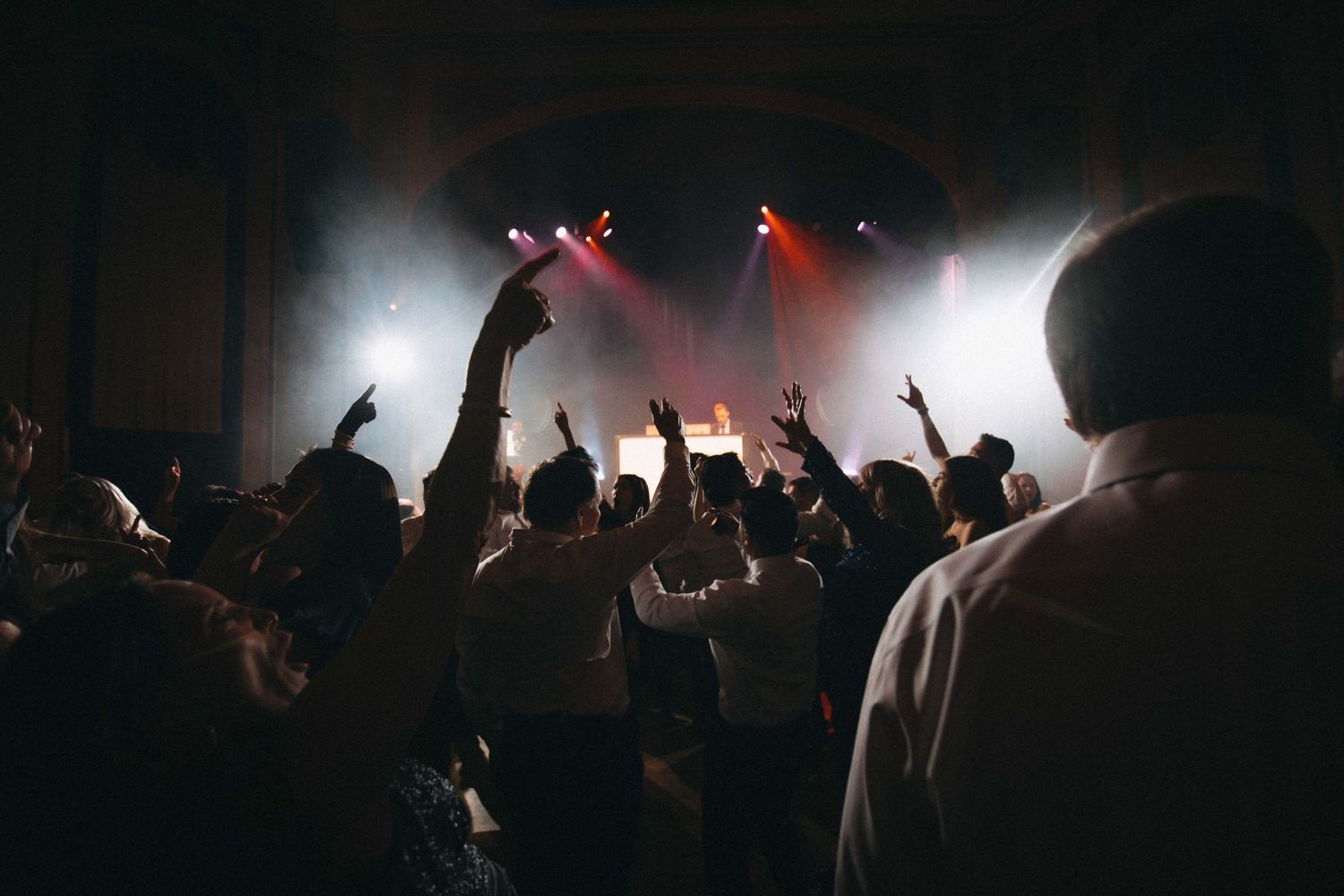 Energetic wedding reception crowd dancing under dramatic stage lights with hands raised.