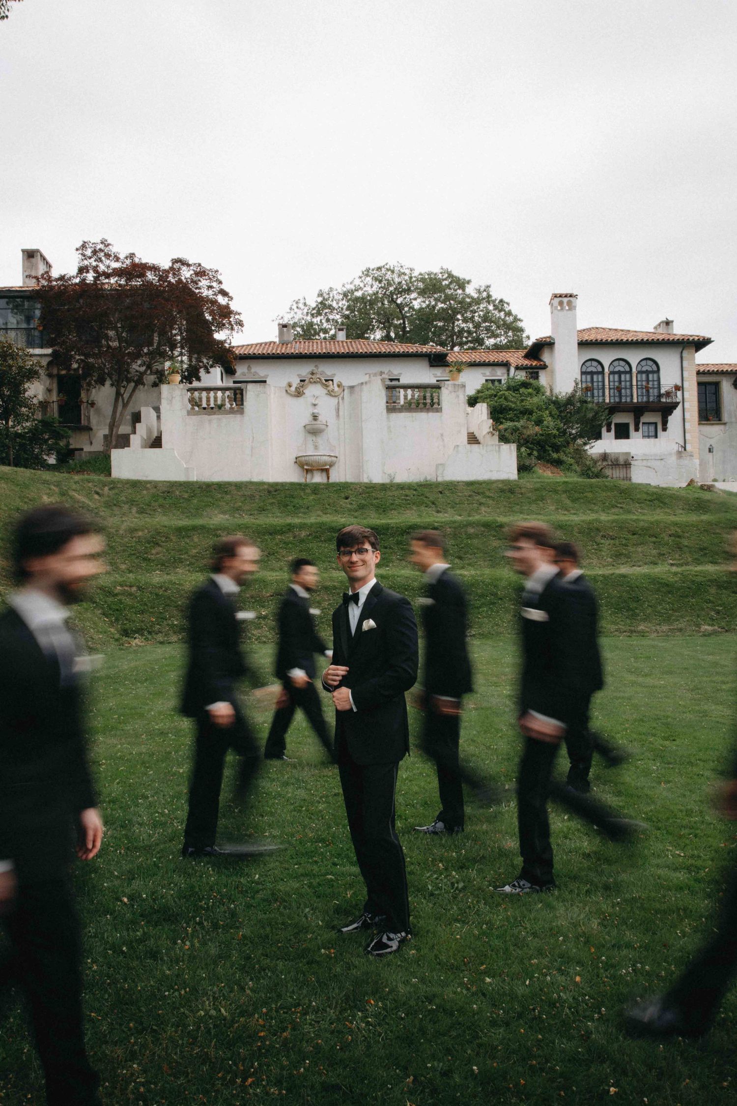Groom stands still in black tuxedo while groomsmen blur past on green lawn before historic mansion.