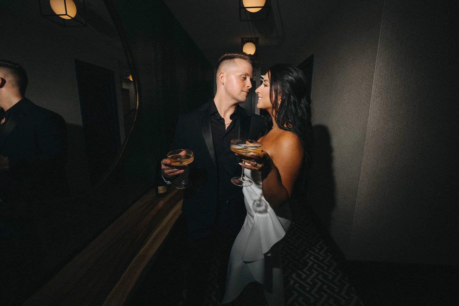 Elegant couple holding cocktails in intimate dark bar setting, woman in black dress, man in suit.