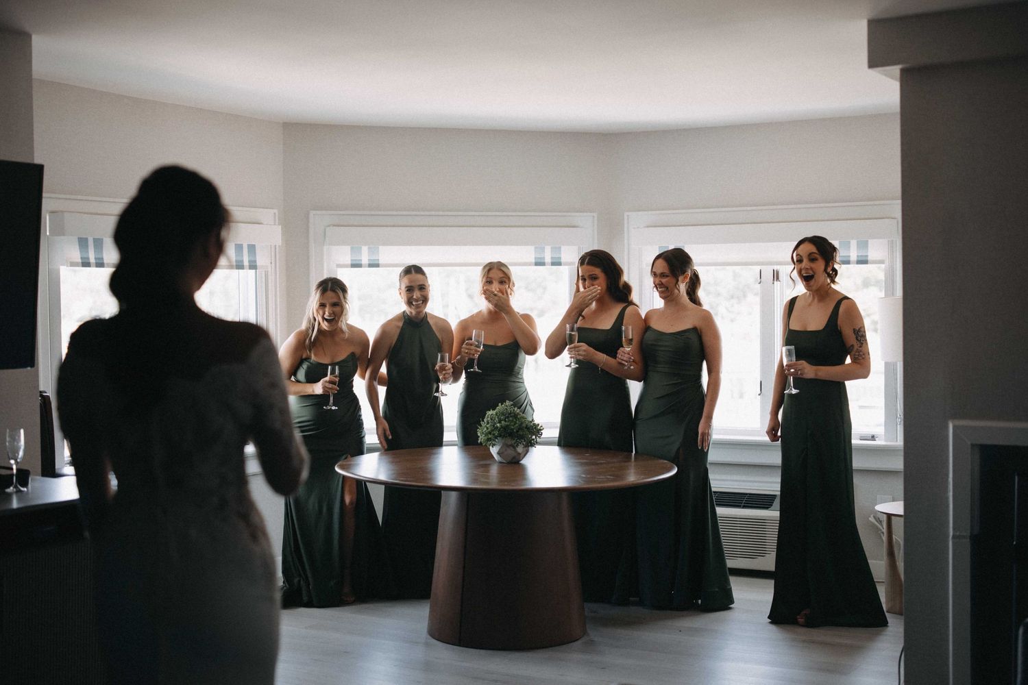 Bridesmaids in dark green gowns react emotionally as bride reveals her wedding look in bright room.