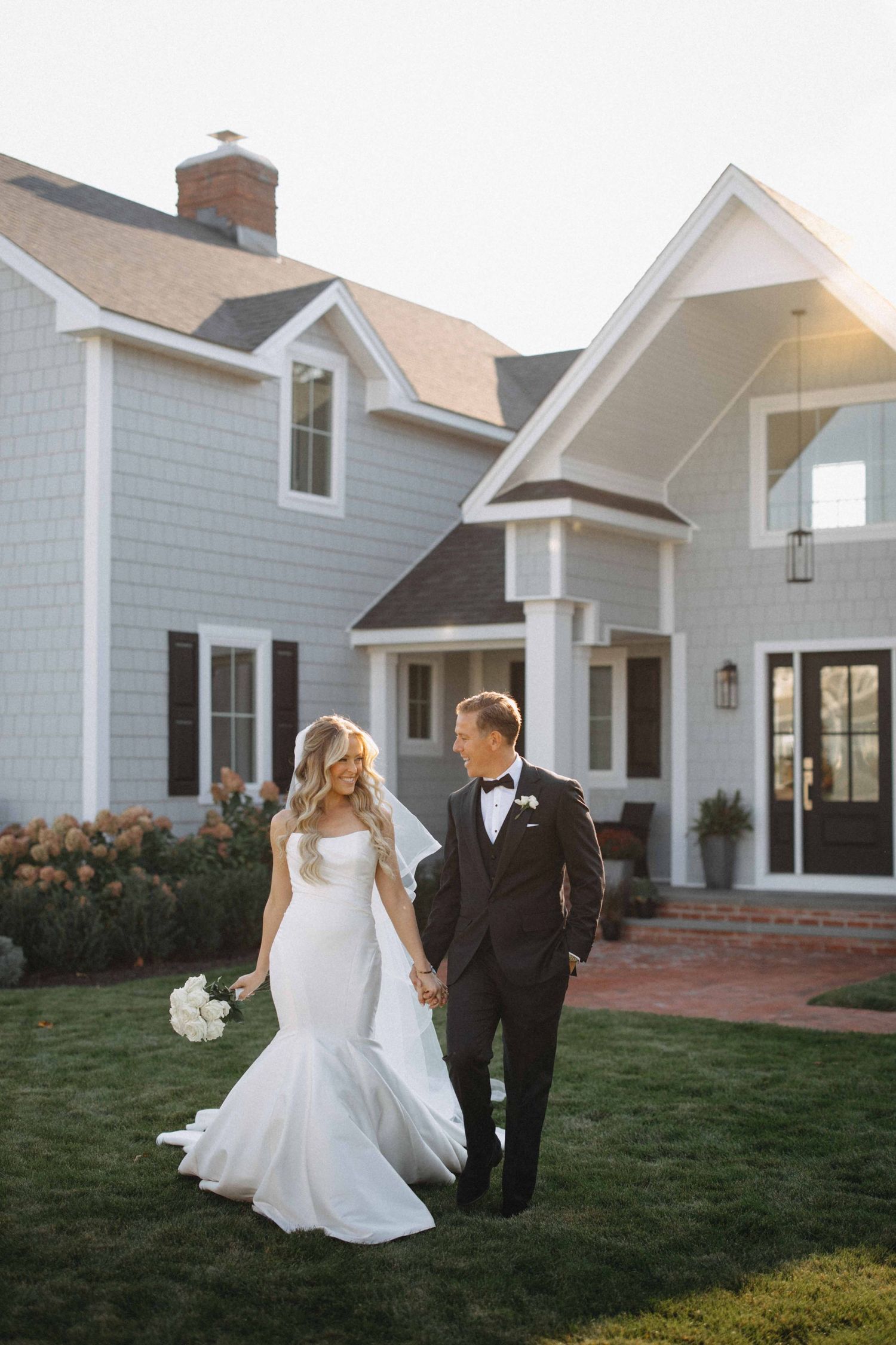 Bride in mermaid gown and groom in tuxedo walk hand-in-hand outside a elegant gray shingle-style estate.